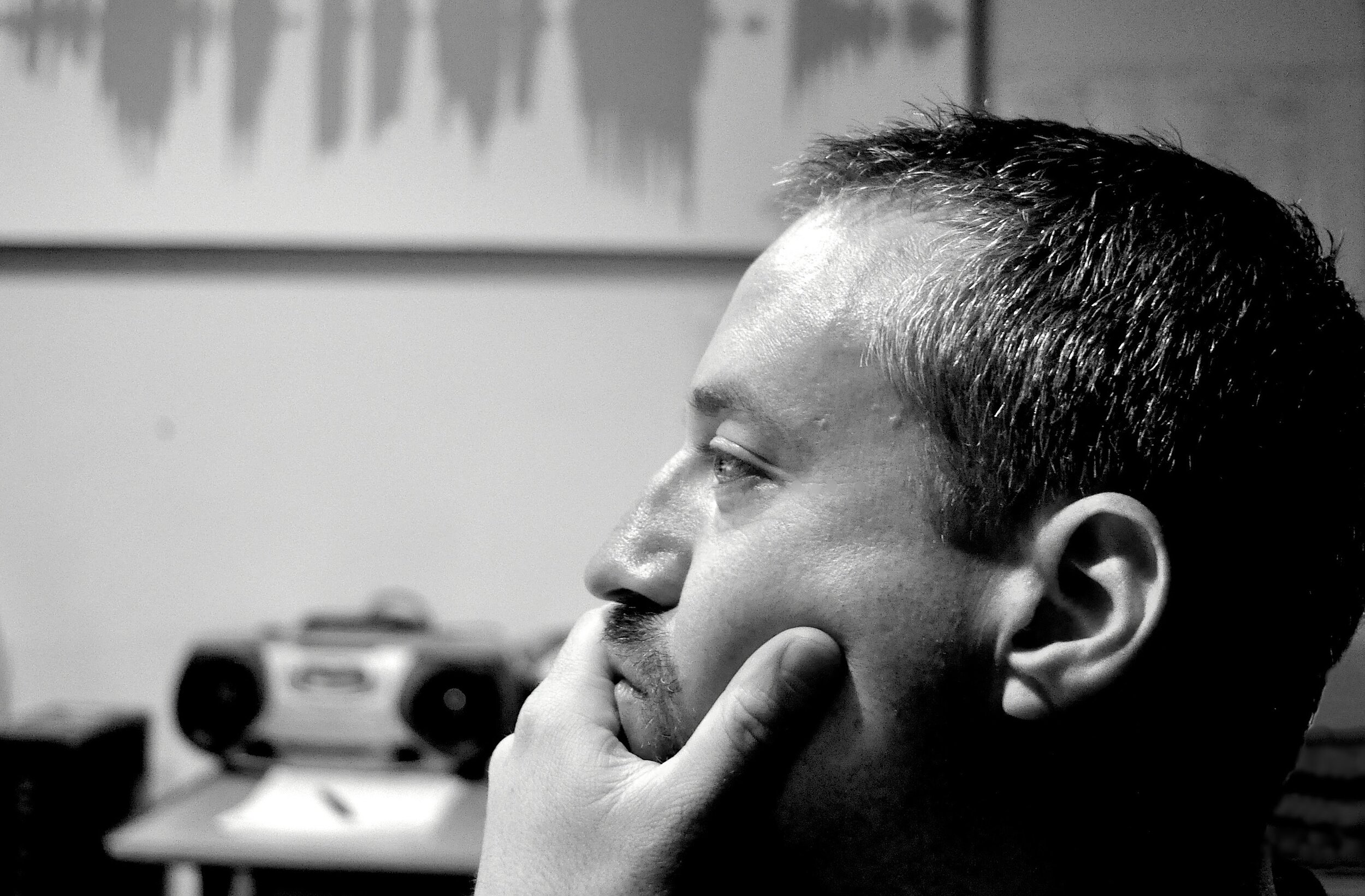 A black-and-white photo of a man in profile, resting his chin on his hand, with a blurred stereo and artwork in the background.