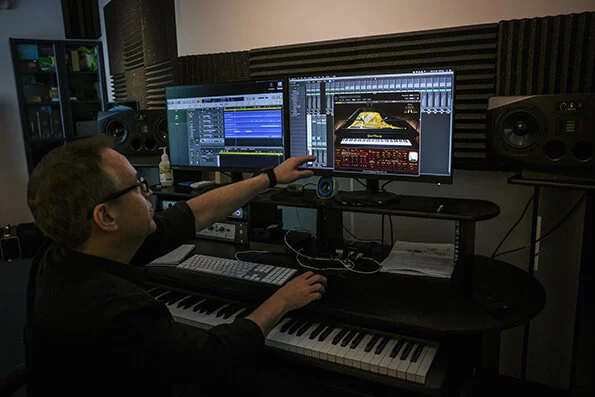 A person working on music production at a computer with multiple monitors displaying audio software, surrounded by audio equipment and soundproofing panels.