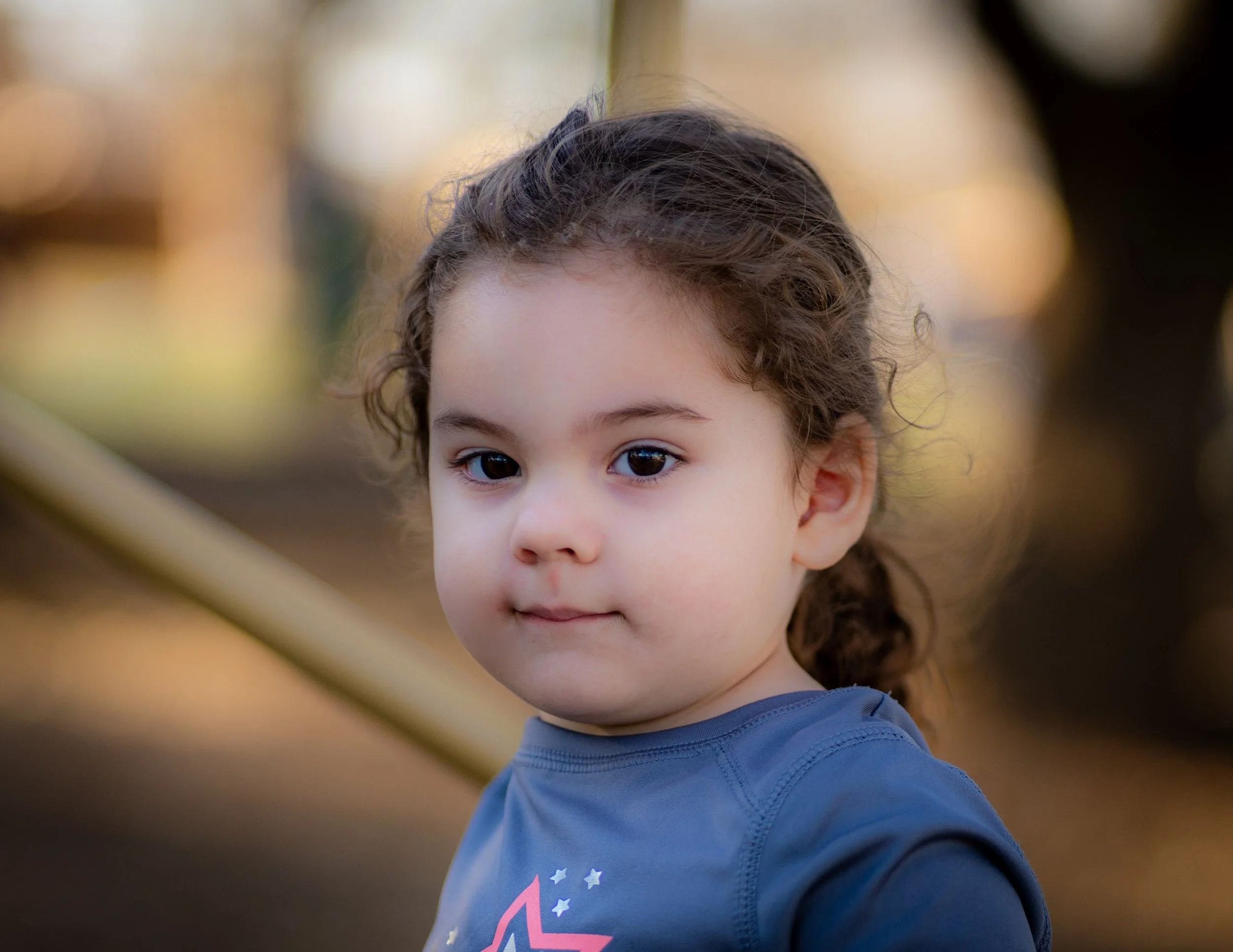 Close-up of a young girl with curly dark hair, wearing a blue shirt, with a blurred natural outdoor background.