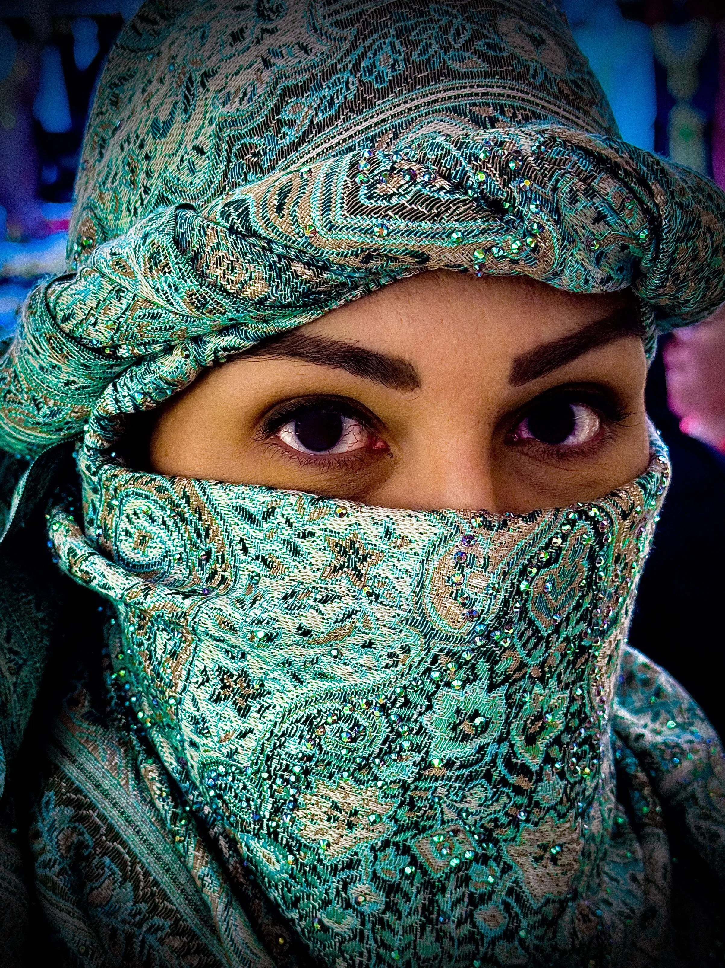 Close-up of a woman's face with only her eyes visible, wearing a patterned, jeweled headscarf that covers her head and lower face.