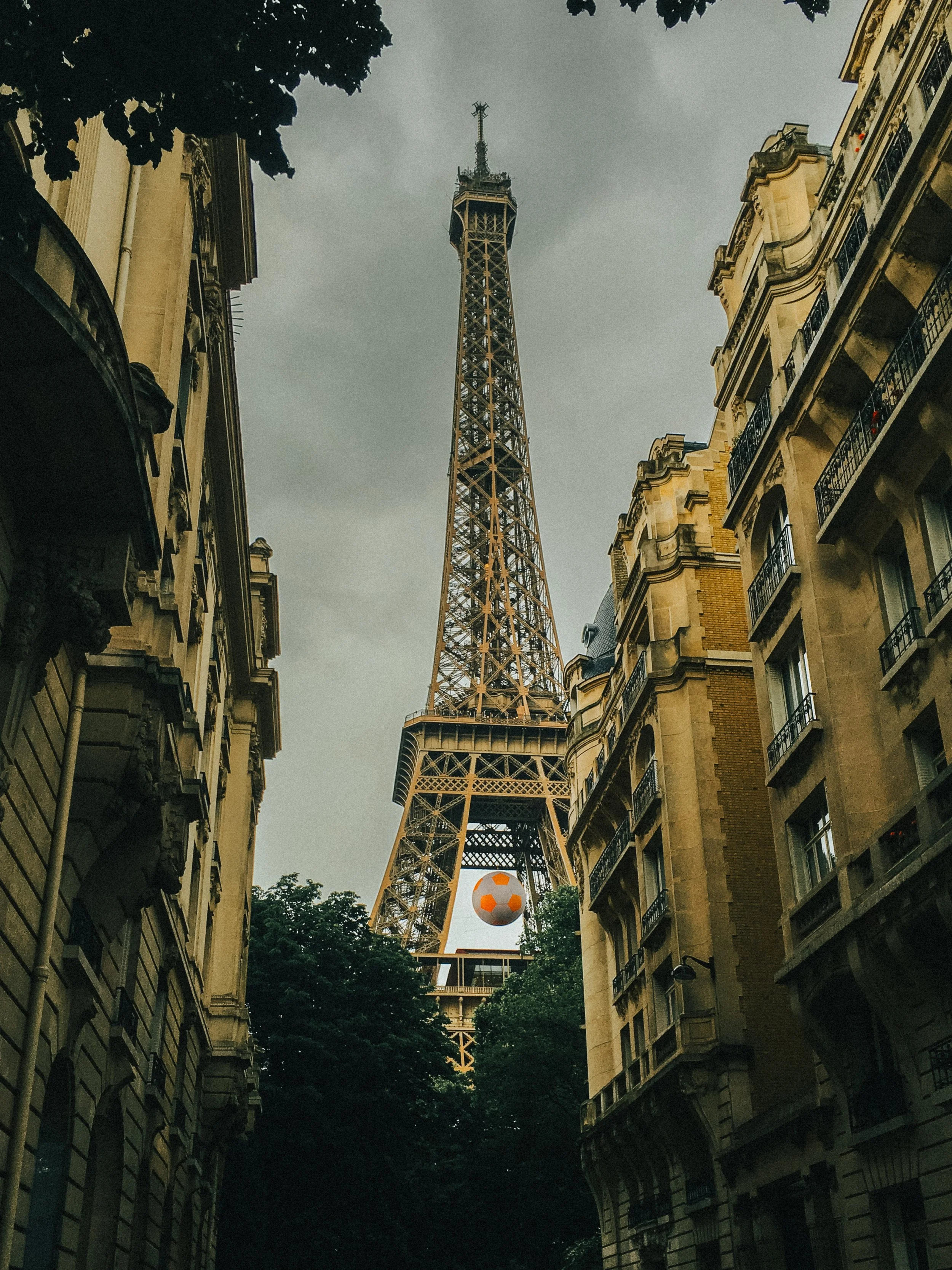 The Eiffel Tower viewed from a narrow street lined with classic Parisian apartment buildings, with a soccer ball suspended in front of the tower.
