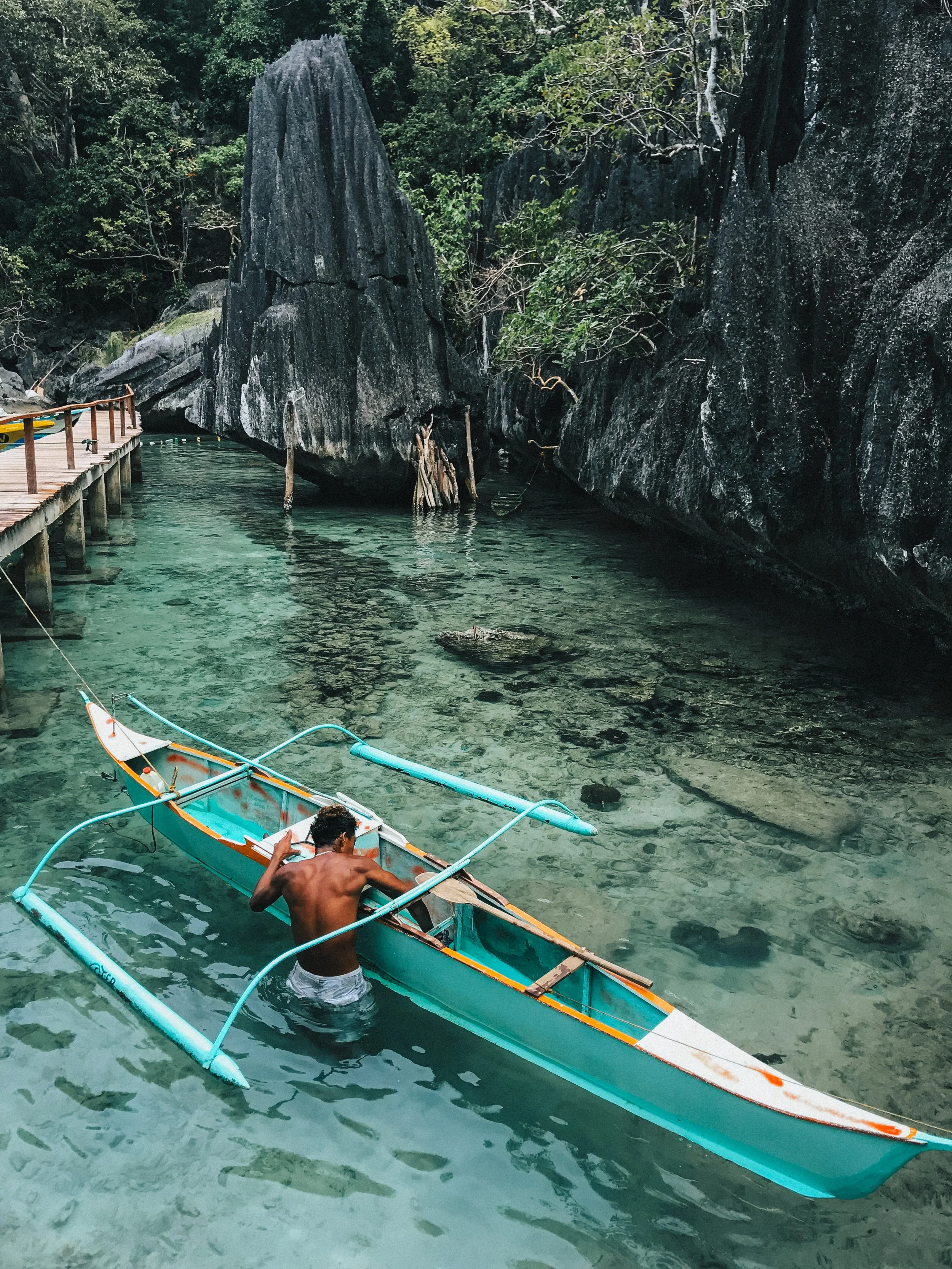 A shirtless man in white shorts standing in clear, shallow water next to a small, colorful boat with outriggers, surrounded by large dark rocks and green trees.