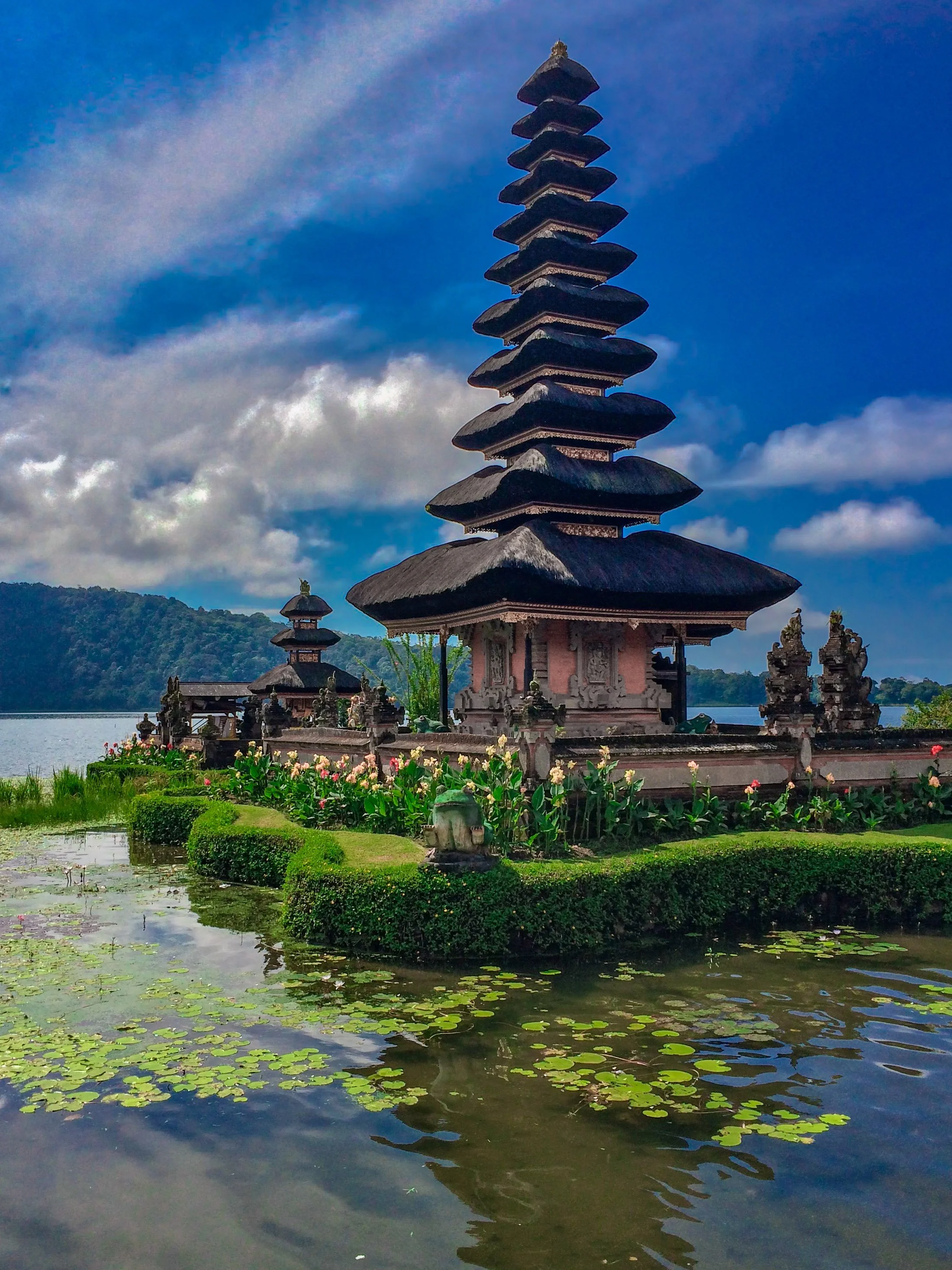 Traditional Balinese temple with multiple tiered pagoda roofs, surrounded by water lilies and lush greenery, set against a lakeside background with hills and a partly cloudy sky.