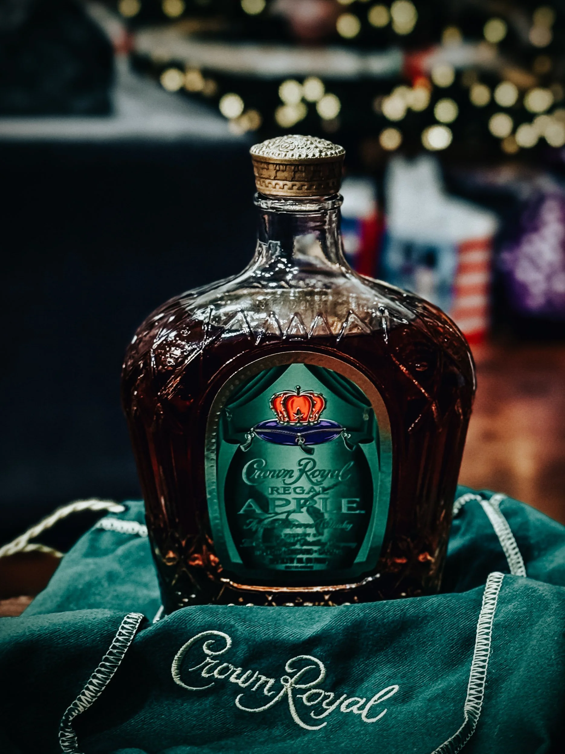 A glass bottle of Crown Royal Apple whiskey with a decorative label and a crown logo, placed on a green cloth with white stitching and embroidered text, with a blurred background of festive holiday decorations.