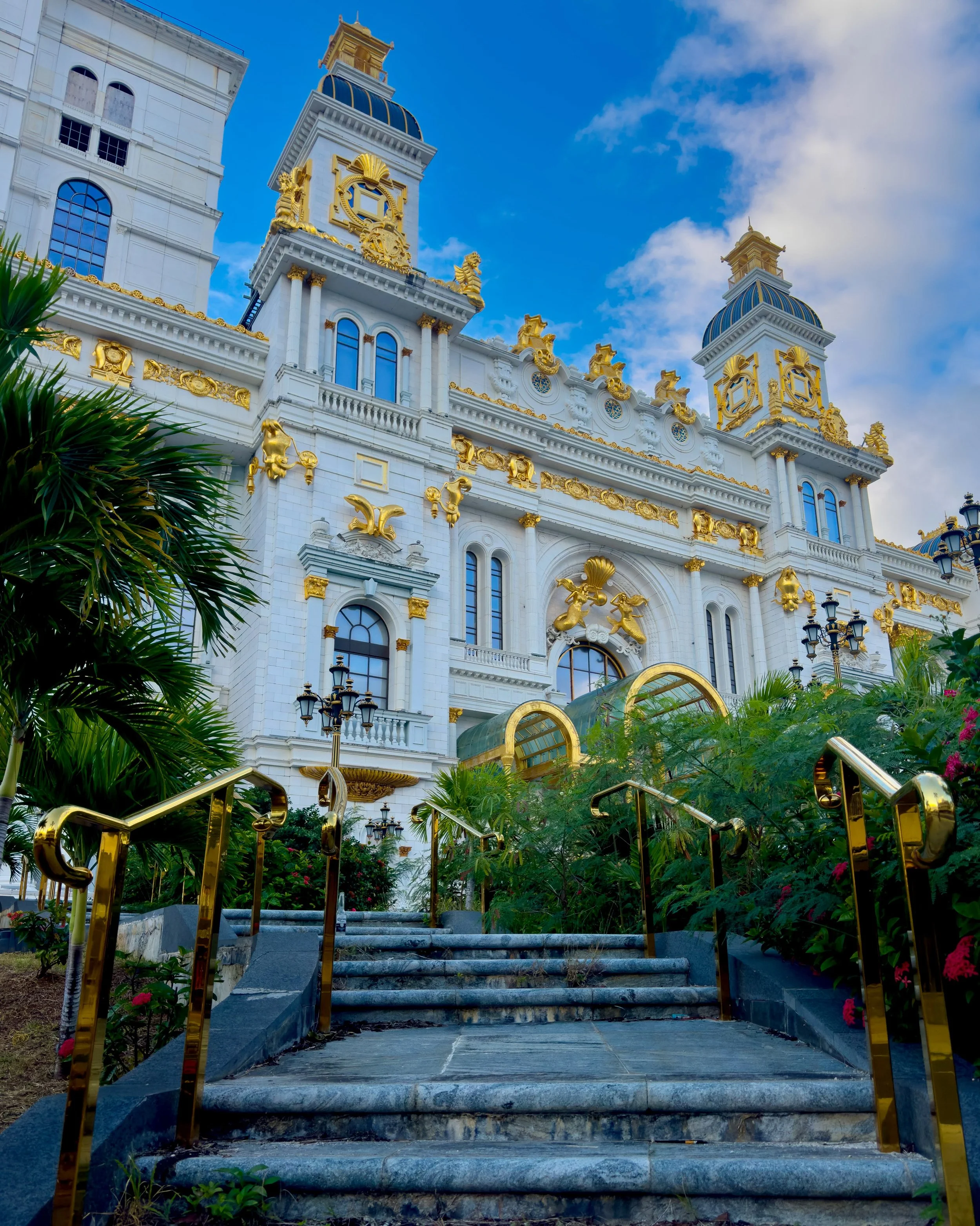 A grand white castle-like structure with gold accents, multiple turrets, and ornate architectural details, surrounded by greenery and manicured plants, under a blue sky with scattered clouds.