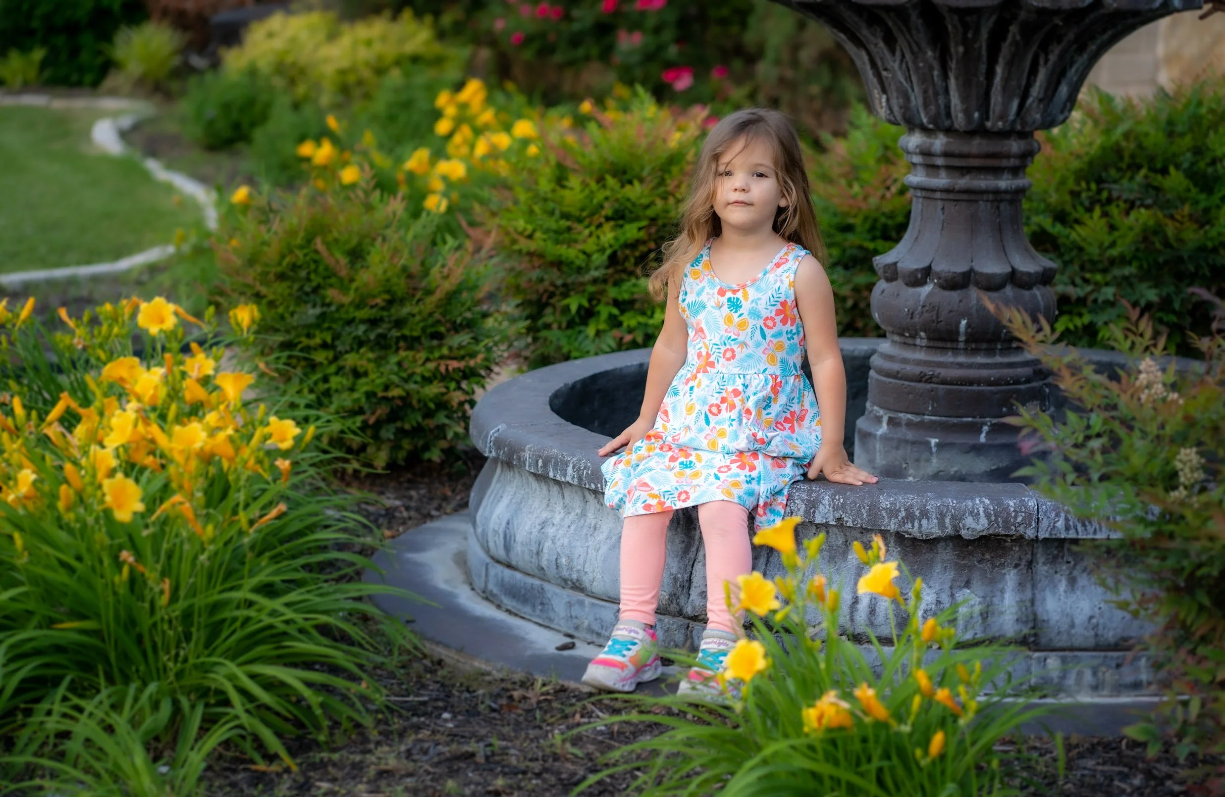 A young girl sitting on the edge of a garden fountain surrounded by yellow flowers and green shrubbery.