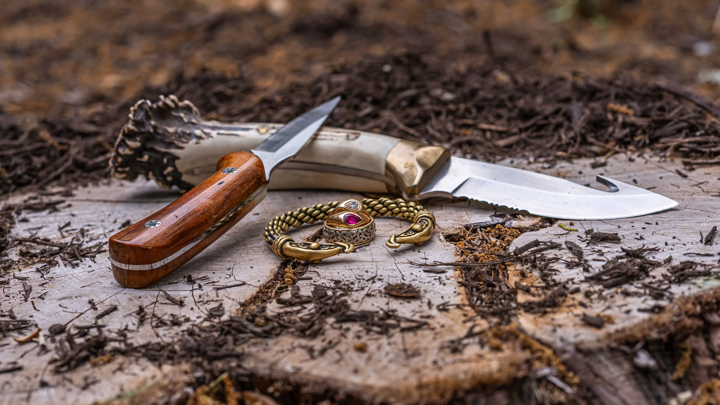 A hunting knife with a wooden handle, a gold jewelry bracelet with a red gemstone, and a gold ring with a clear gemstone placed on a tree stump among scattered wood chips and dirt.