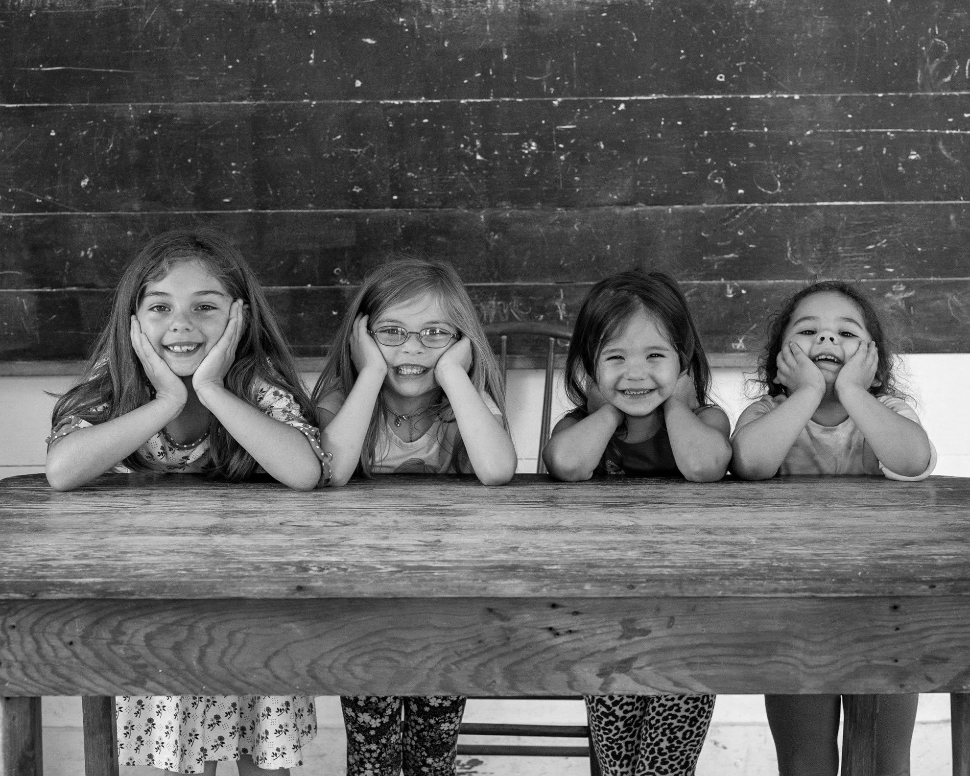 Five young girls sitting at a wooden table, resting their heads on their hands, smiling and posing in front of a chalkboard.