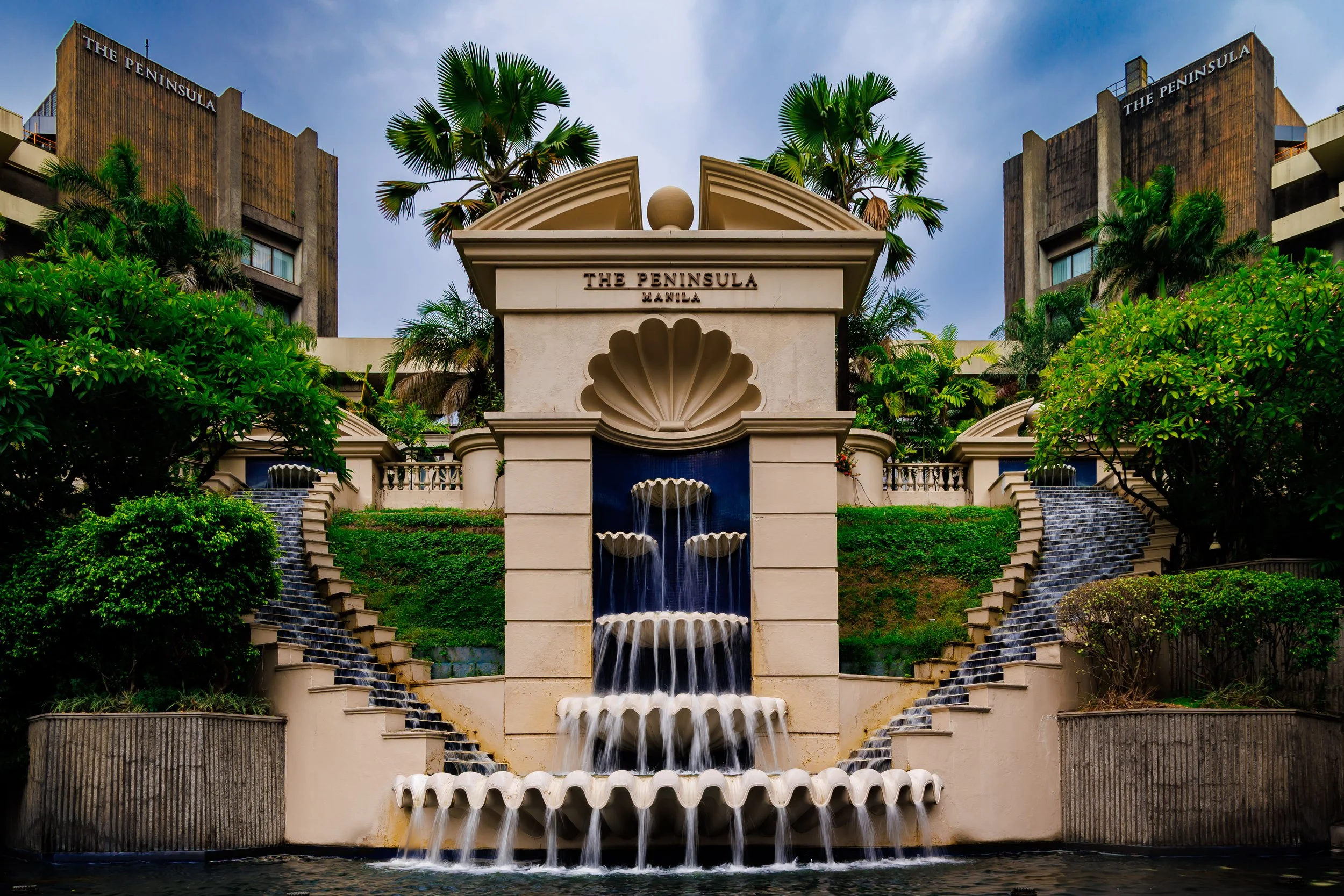 A decorative fountain with cascading water in front of a beige structure labeled 'The Peninsula Manila,' with lush greenery and palm trees, and two tall buildings in the background.