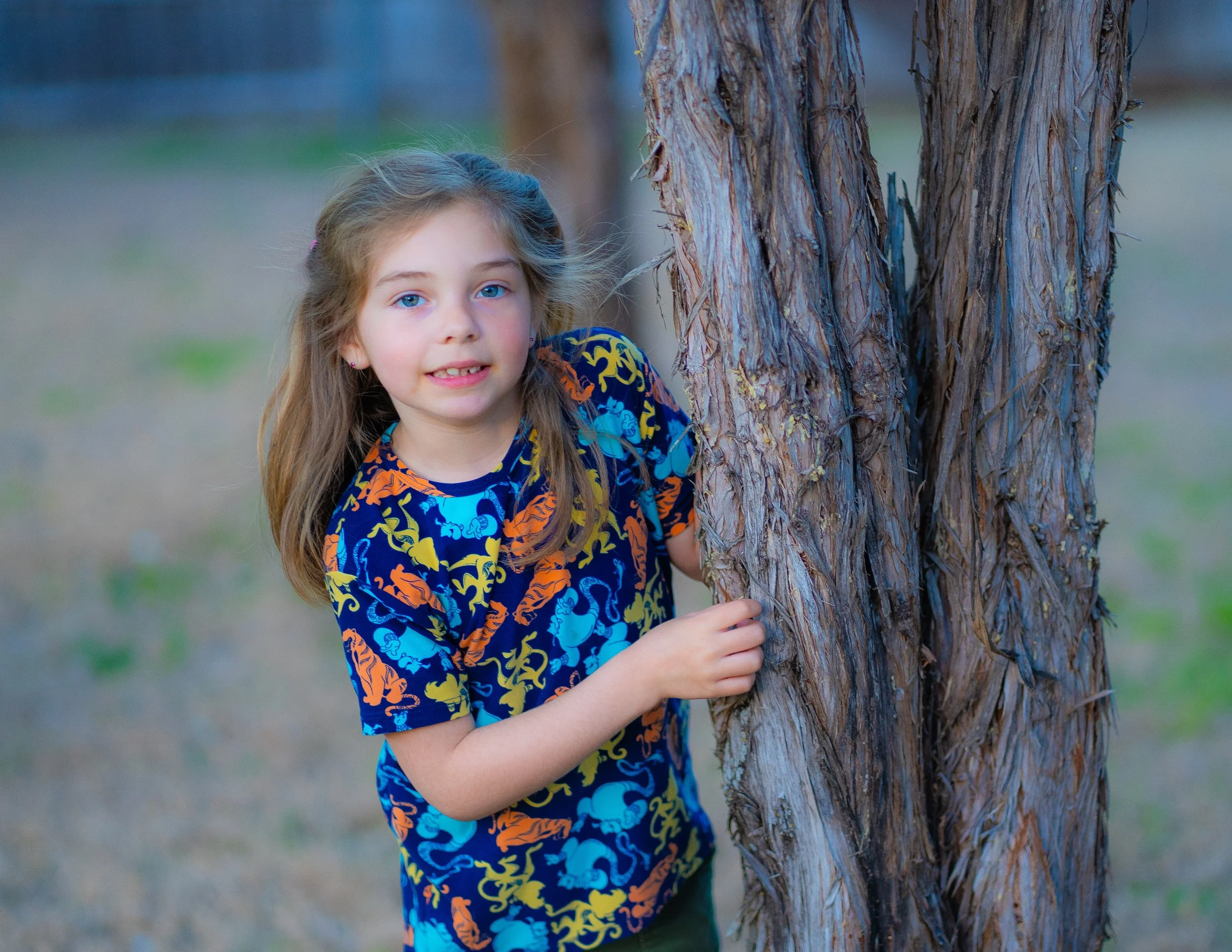 A young girl with long light brown hair and blue eyes peeking from behind a tree in a park.