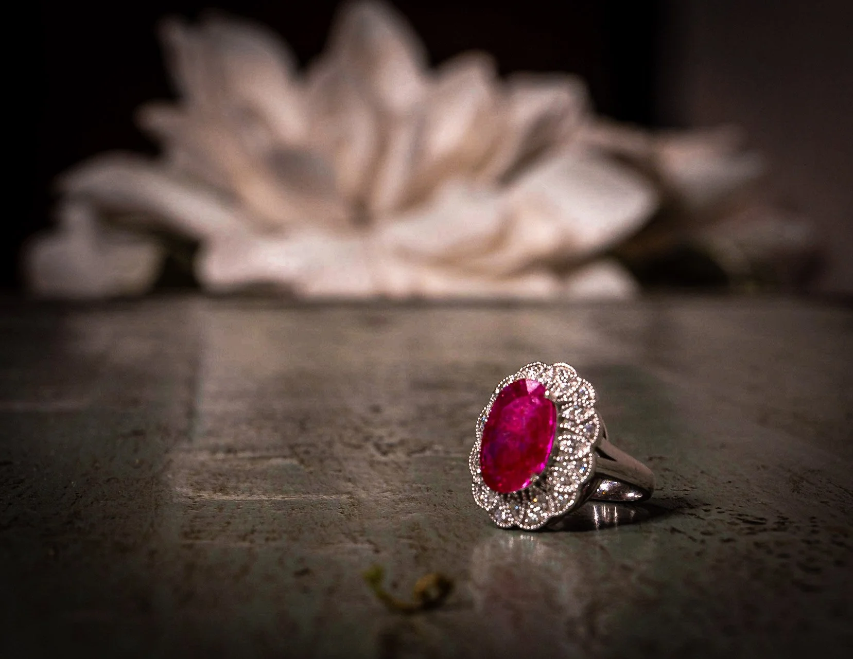 A silver ring with a large, oval-shaped ruby surrounded by small diamonds on a wooden surface, with a blurred light-colored flower in the background.