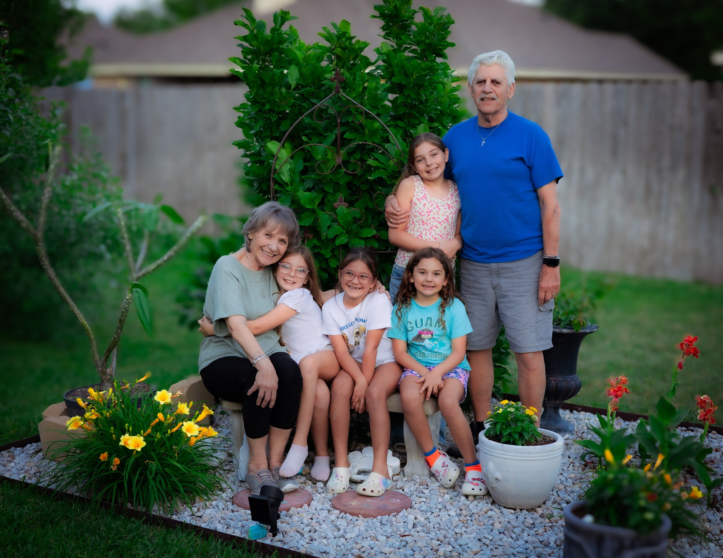 Family photo in a backyard garden with an elderly woman, four young girls, and an elderly man, surrounded by potted flowers, a green shrub, and a wooden fence.