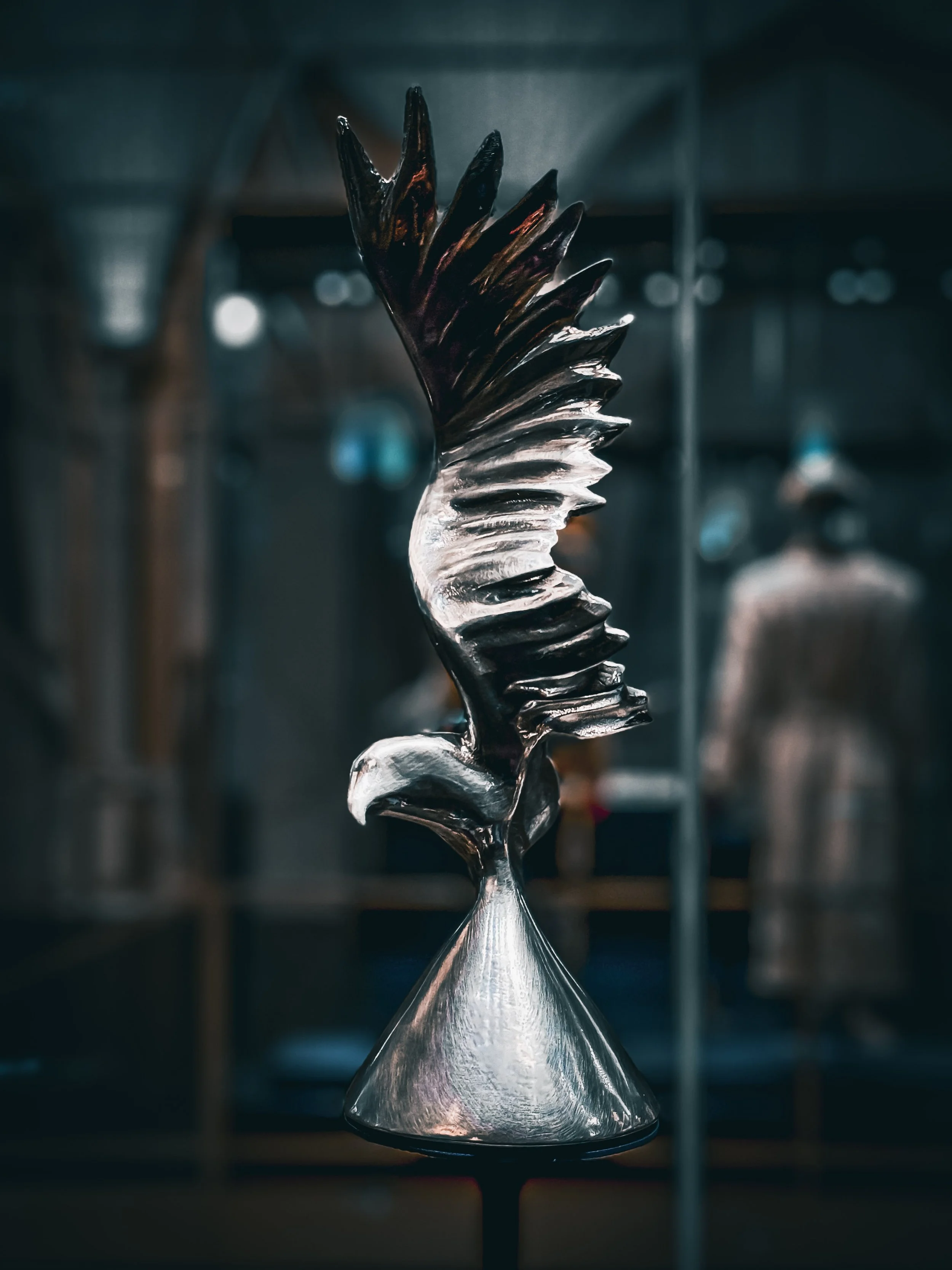 A metallic sculpture of an eagle with wings spread, mounted on a conical base, displayed in a gallery or exhibit setting.
