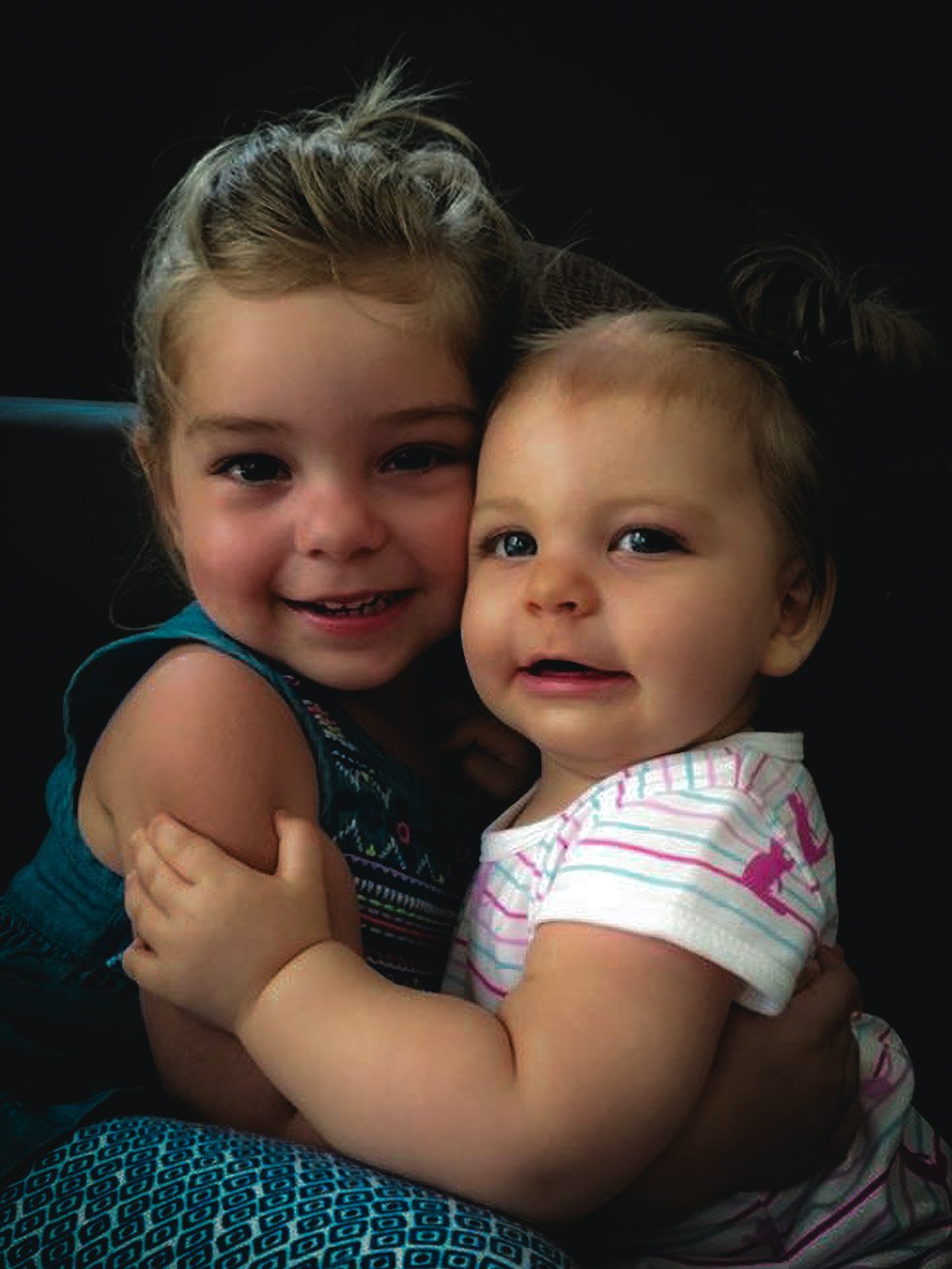 Two young girls hugging and smiling, with a dark background.