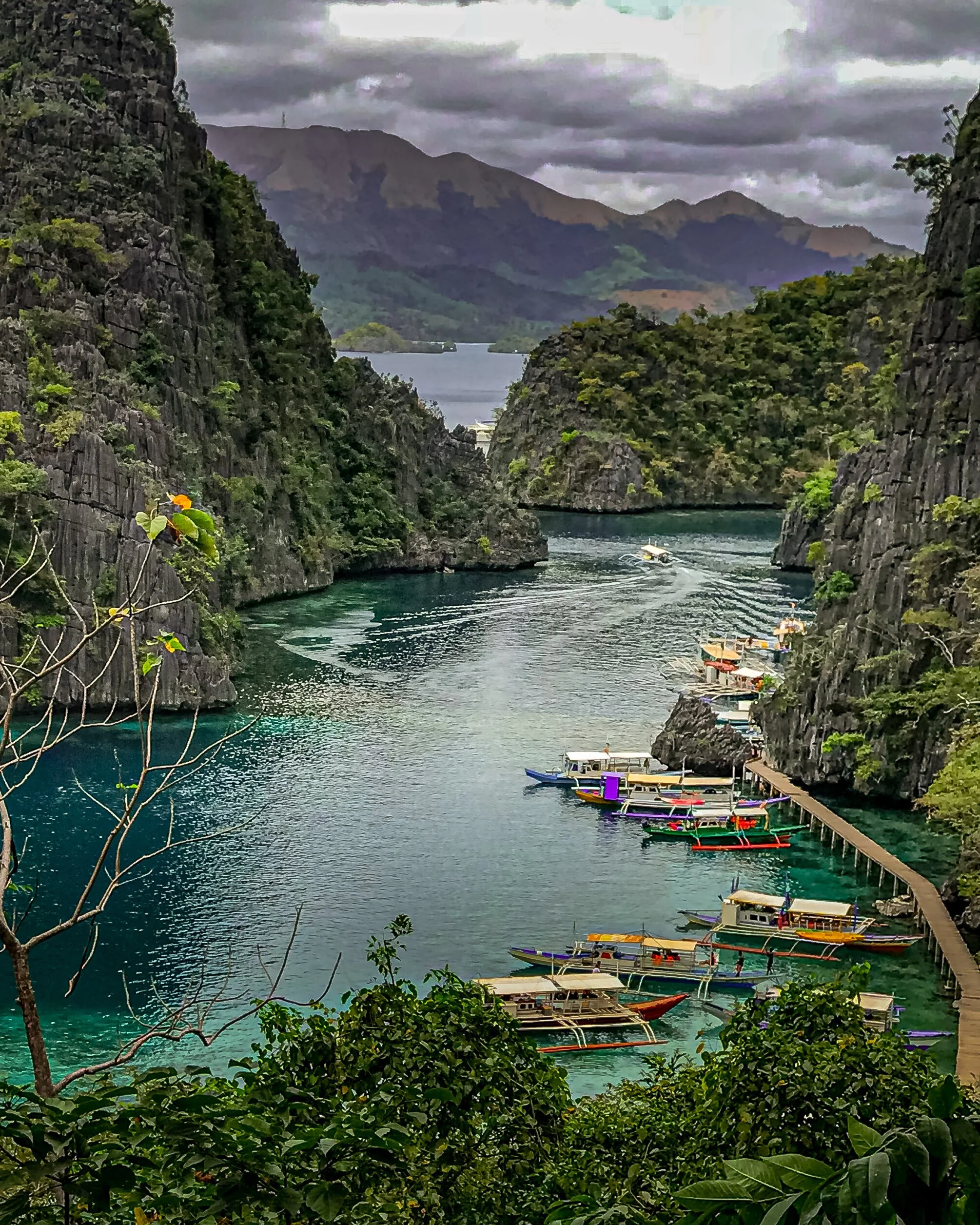 Tropical island canyon with steep green cliffs, calm turquoise water, boats docked along a pier, and mountains with cloudy sky in the background.