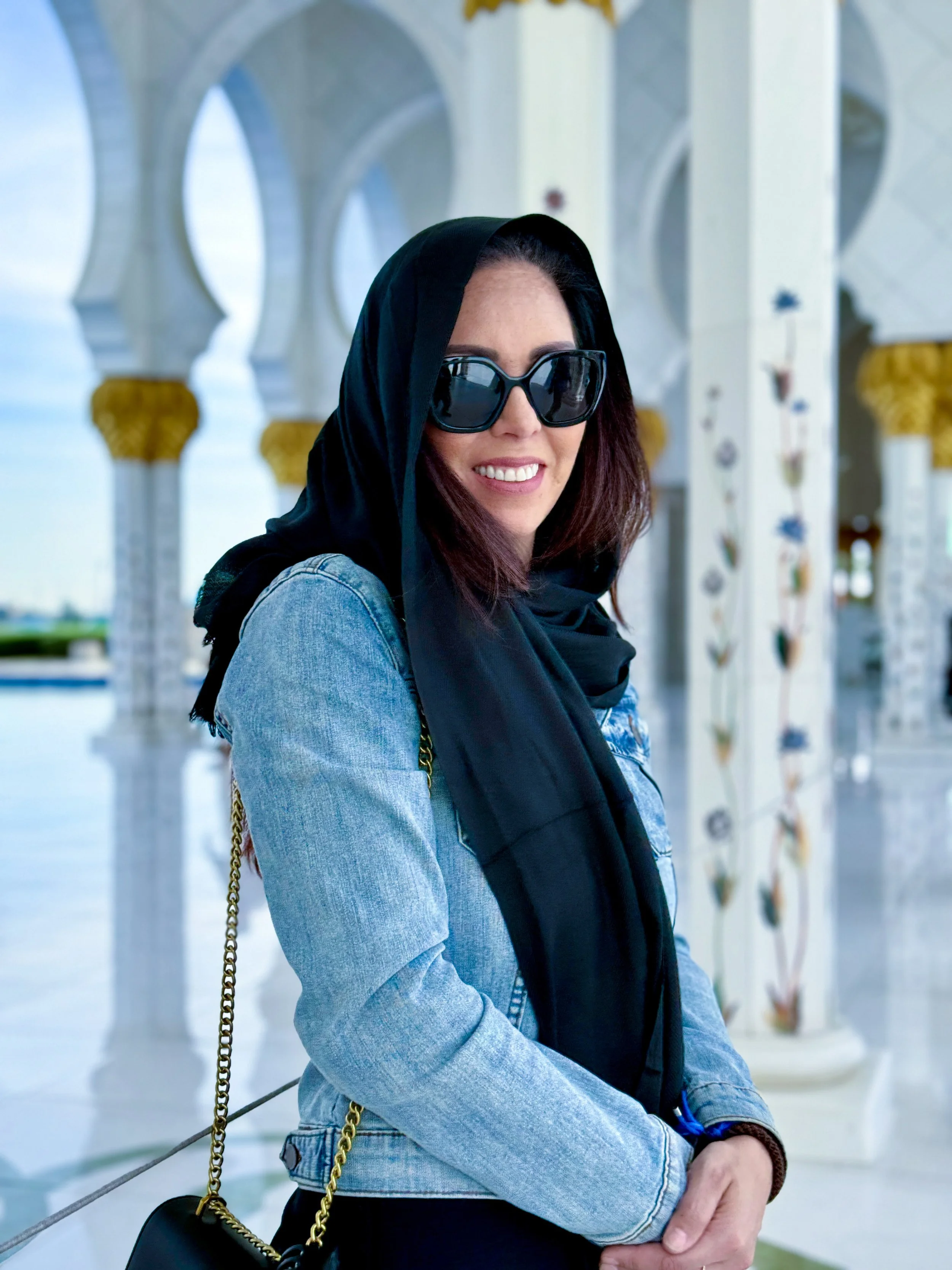 Woman with sunglasses and a black headscarf standing outdoors near ornate white columns with gold accents.