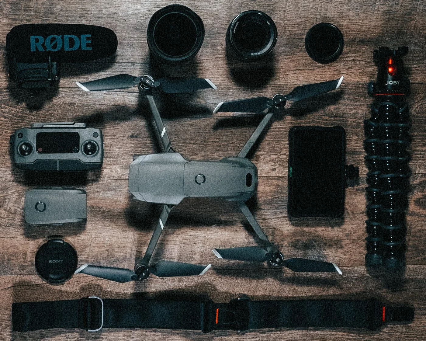 A drone with three propellers, a camera, a controller, a smartphone, a lens, a tripod, a strap, and other camera accessories arranged on a wooden surface.