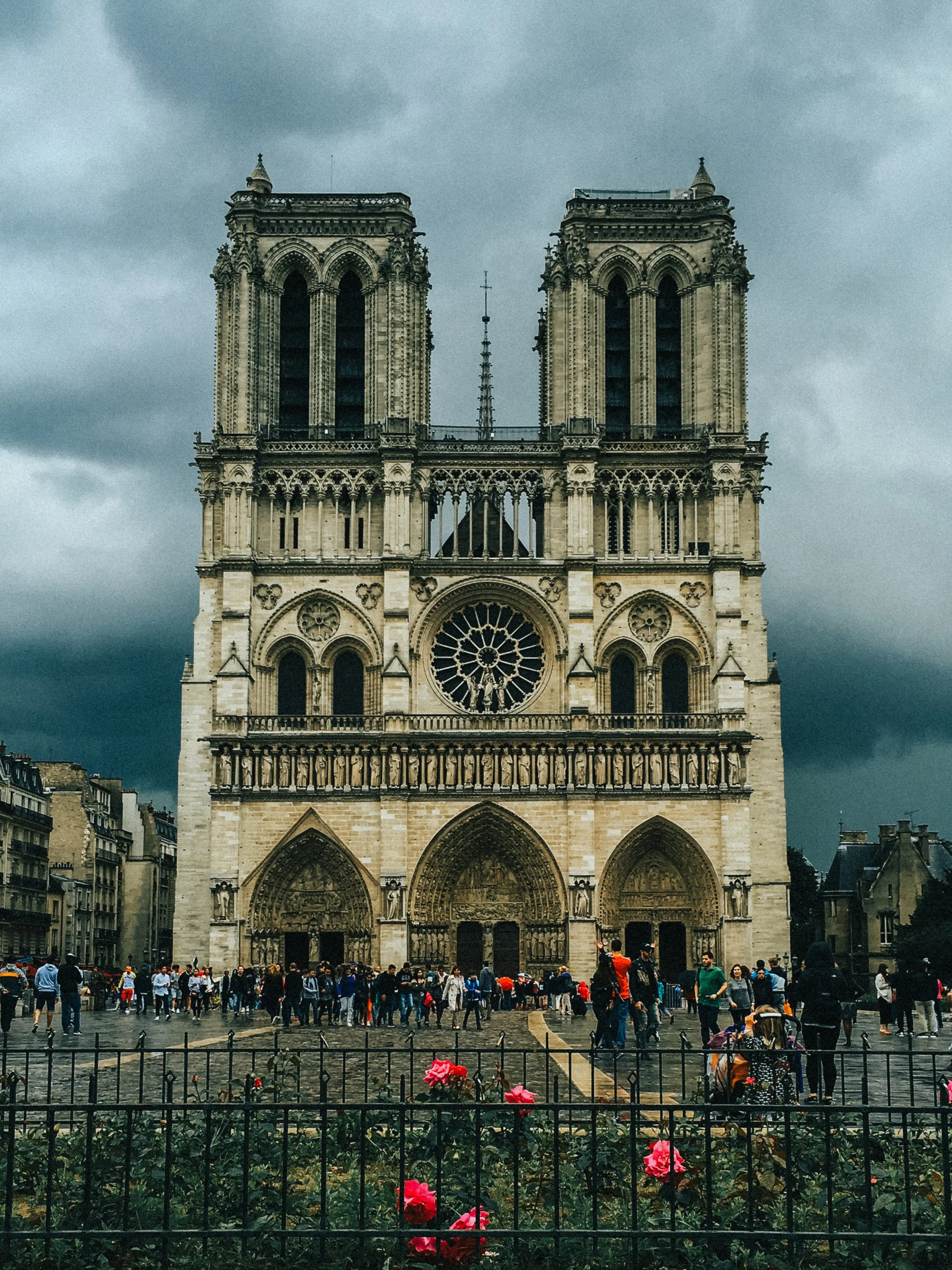 Front view of Notre-Dame Cathedral in Paris under dark, cloudy sky, with many visitors in the plaza and pink roses in the foreground.