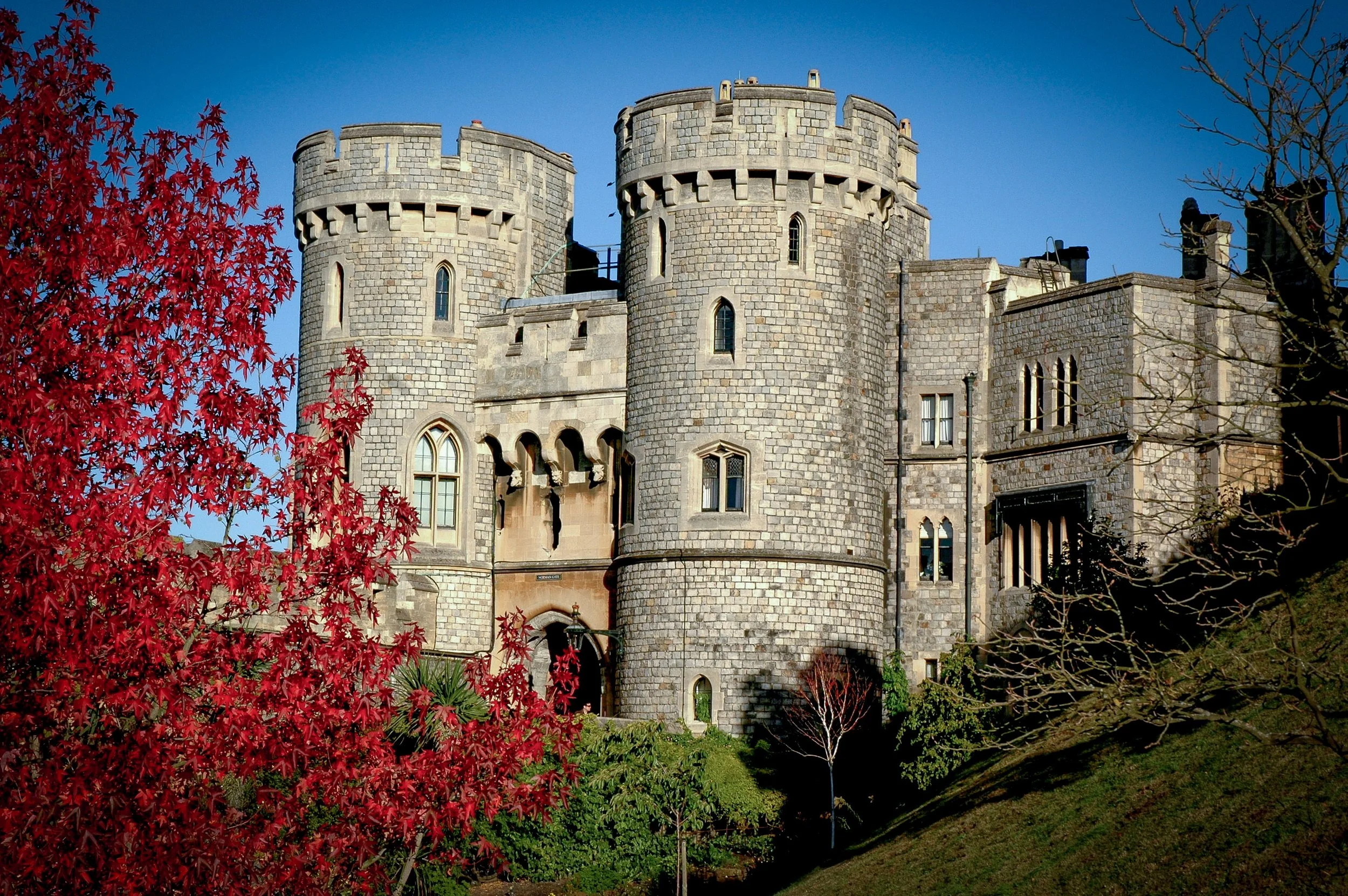 A castle with towers made of gray stone, surrounded by trees with red and brown leaves, under a clear blue sky.