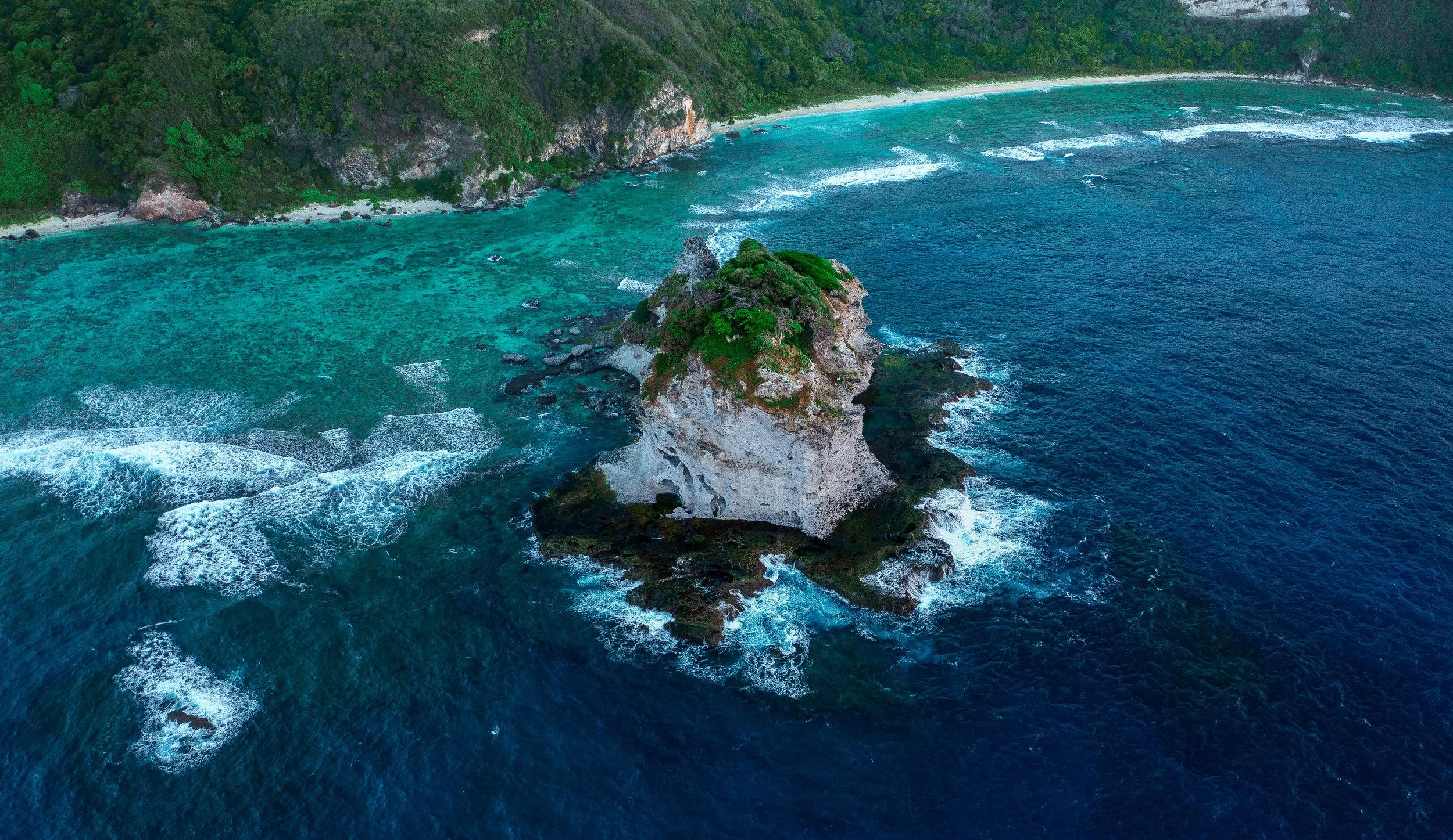 An aerial view of a large rocky island with green vegetation, surrounded by deep blue ocean and waves, and a lush green coastline with sandy beach in the background.