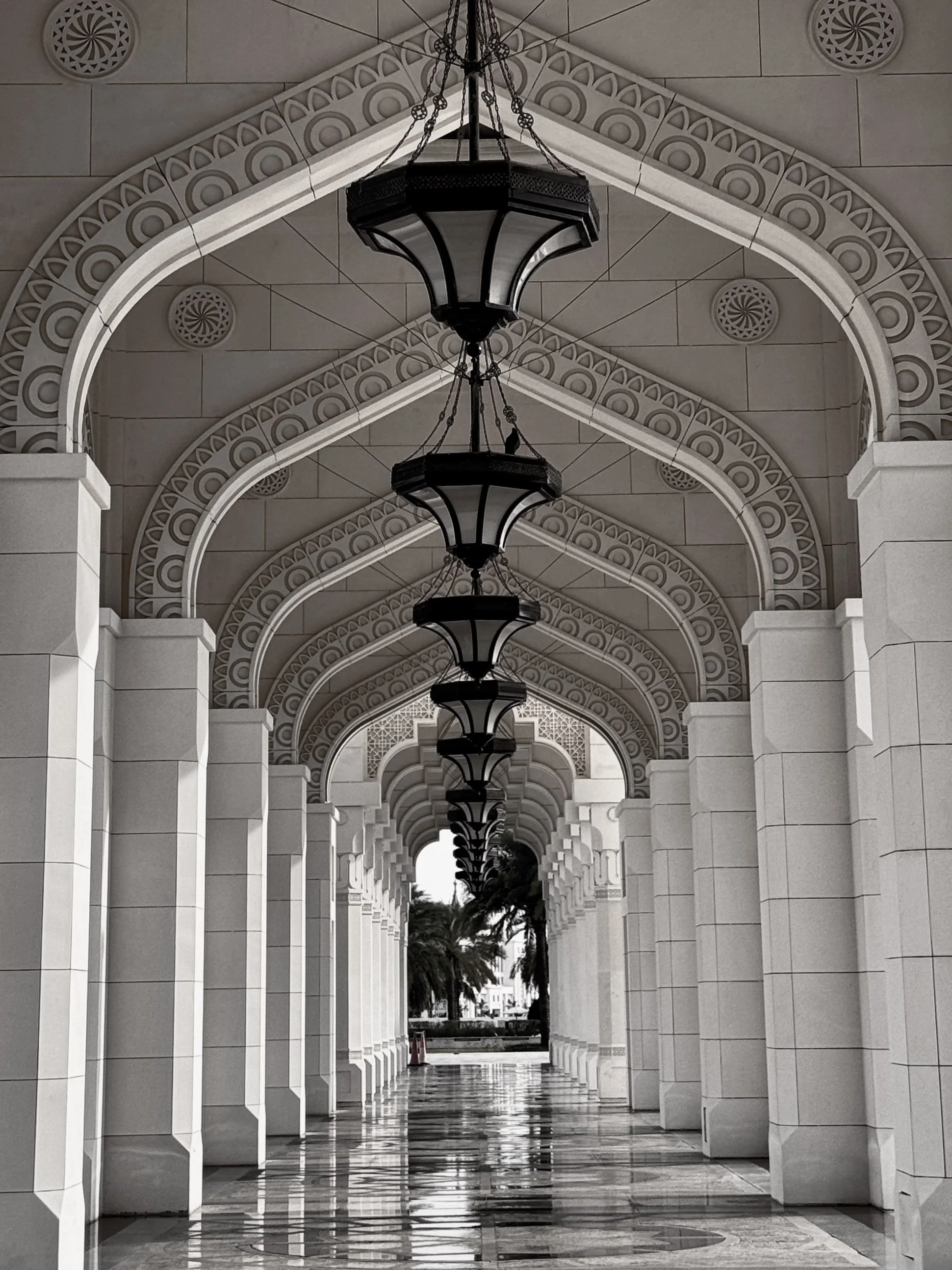 A black and white photo of a long corridor with arches, decorative ceiling, and hanging lantern-style lights. Palm trees are visible at the end of the corridor.