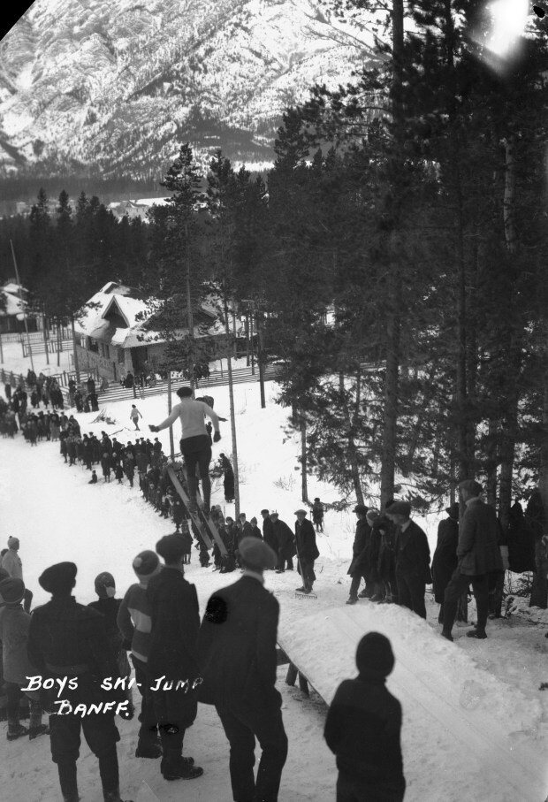   Banff Winter Carnival, boys ski jump, Wolverine Street. We recommend you wander up there (to the base of the Ken Madsen Trail in Banff, Alberta) and see for yourself if the clear-cut is still visible today.    Created by Byron Harmon, 1920. Courtes