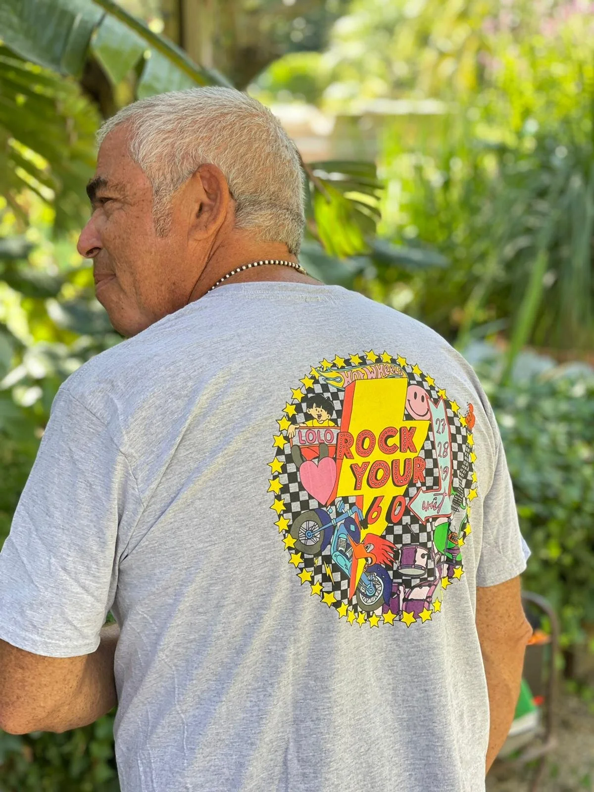 Un homme vu de dos portant un t-shirt gris avec un graphisme coloré à l'arrière, dans un environnement naturel avec de la végétation verte.