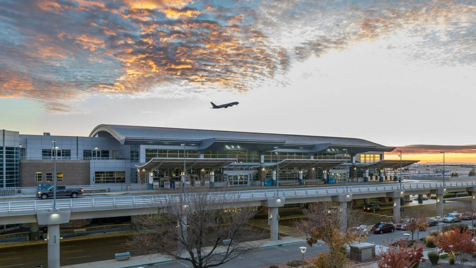 Exterior view of modern airport terminal building at sunset with aircraft in sky and elevated roadway access.