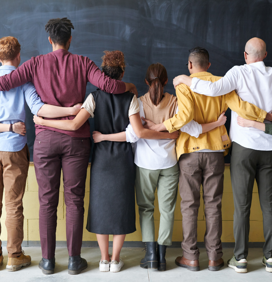 Group of six diverse students standing with arms around each other in front of a chalkboard in a classroom.