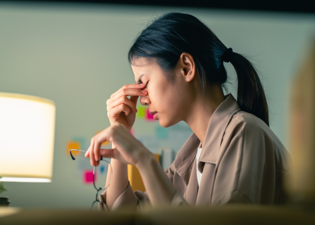 A woman sitting at a desk holding her glasses and pinching the bridge of her nose with a stressed expression.