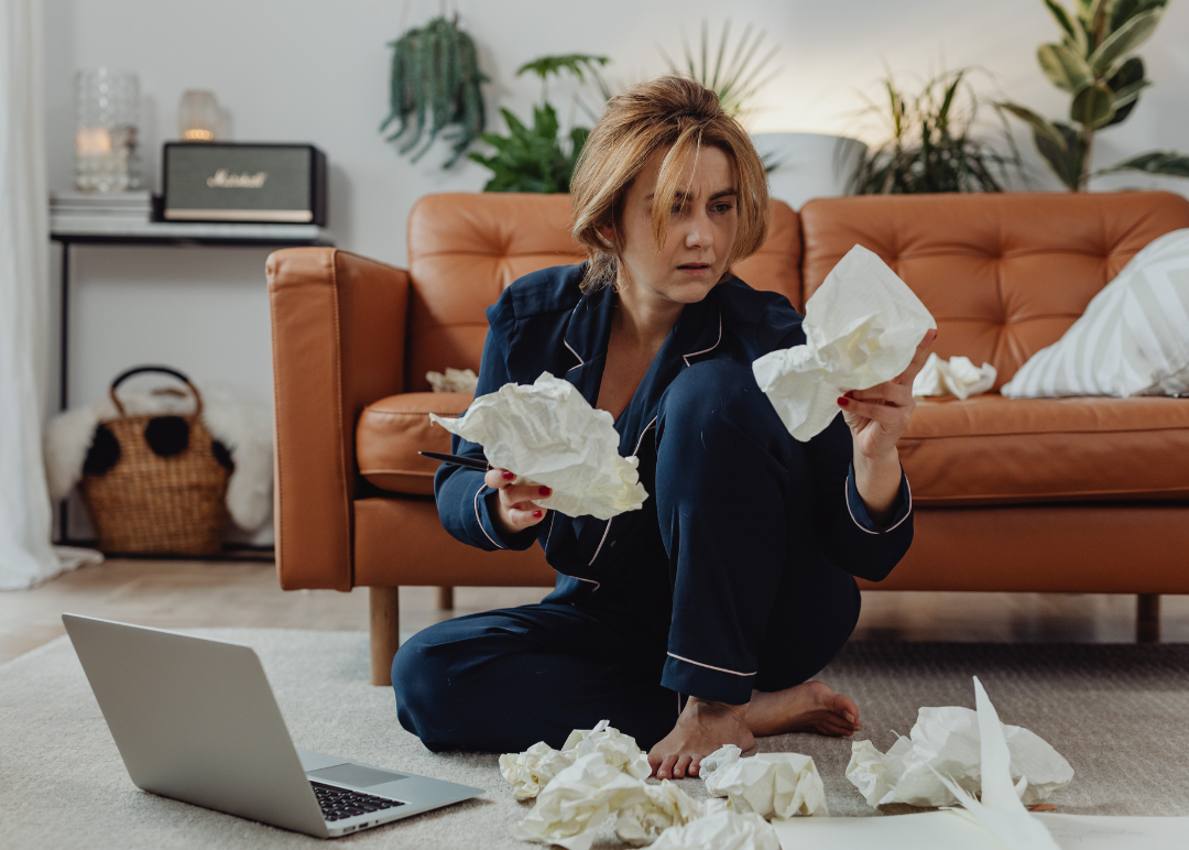 A woman with short blonde hair wearing navy pajamas, sitting on the floor in front of a laptop, surrounded by crumpled tissues, looking distressed or frustrated.