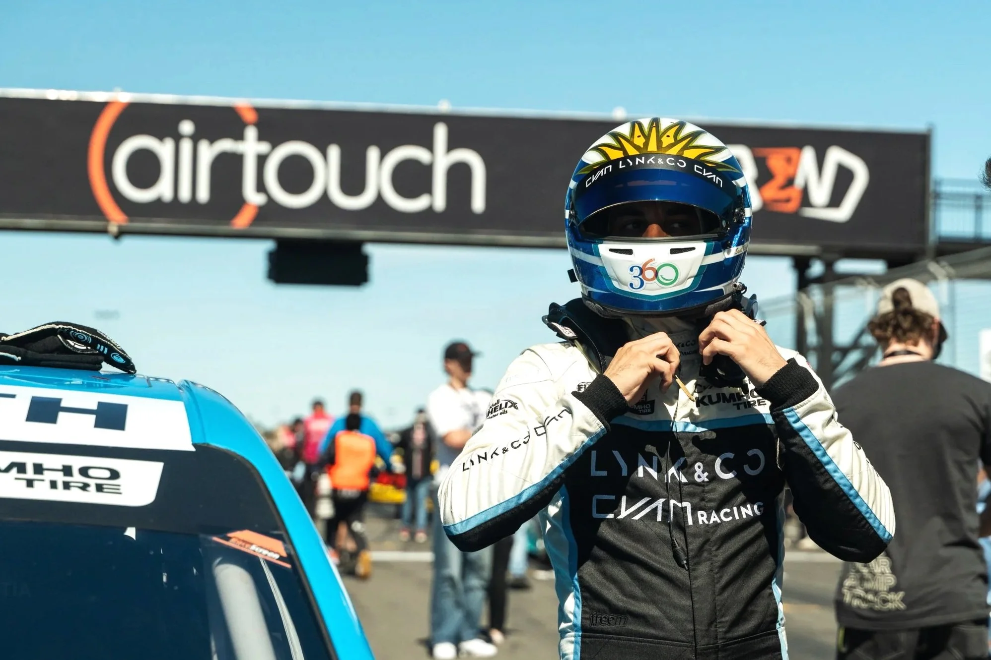 Race car driver in racing suit and helmet adjusting their gear at a race track, with a blue race car and a banner that reads 'airtouch' in the background.