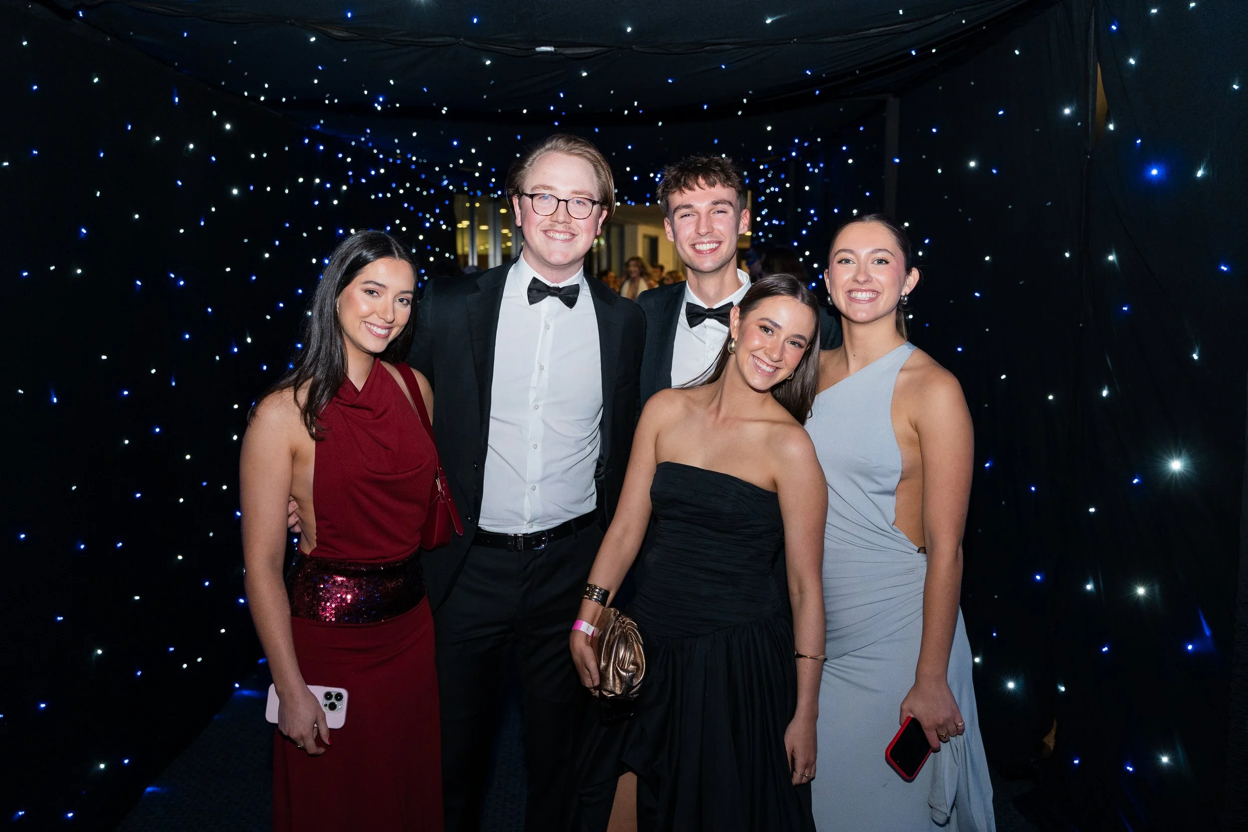 Group of five young adults dressed in formal attire, posing and smiling at an event with a starry, illuminated backdrop.