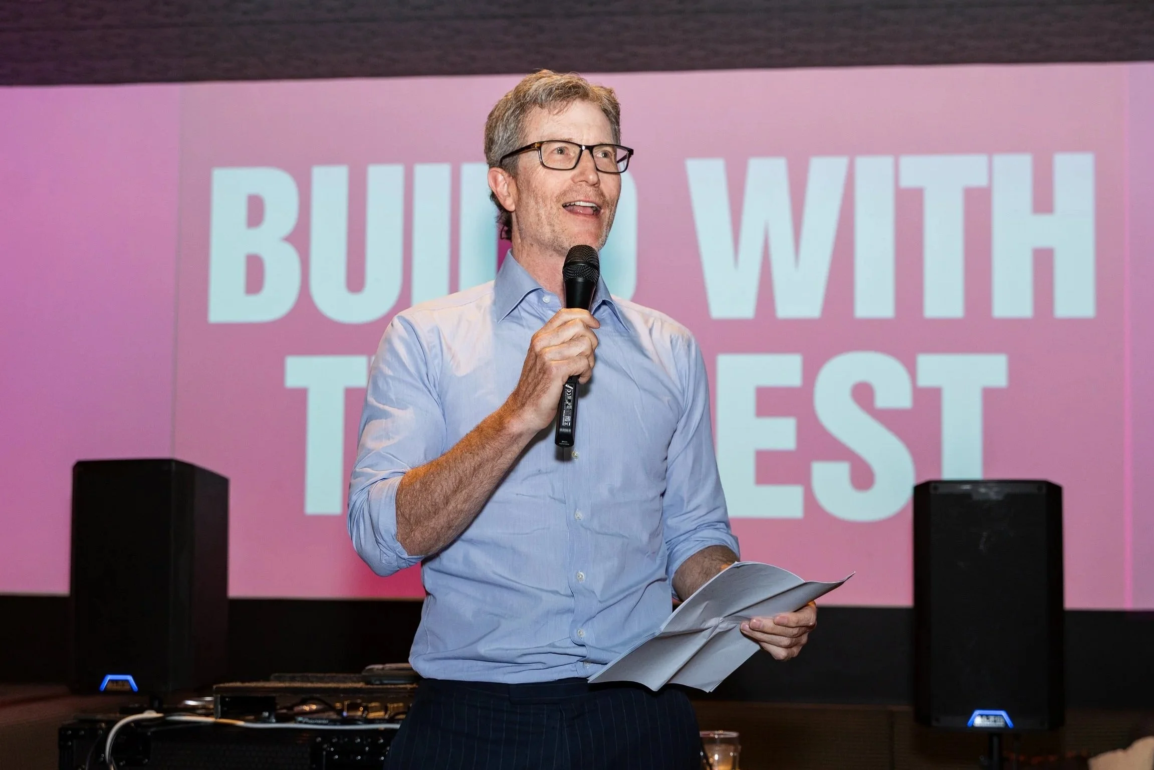 A man with glasses, wearing a light blue shirt, speaking into a microphone while holding papers. Behind him, a pink screen displays the words 'BUILD WITH THE ' in large white letters.