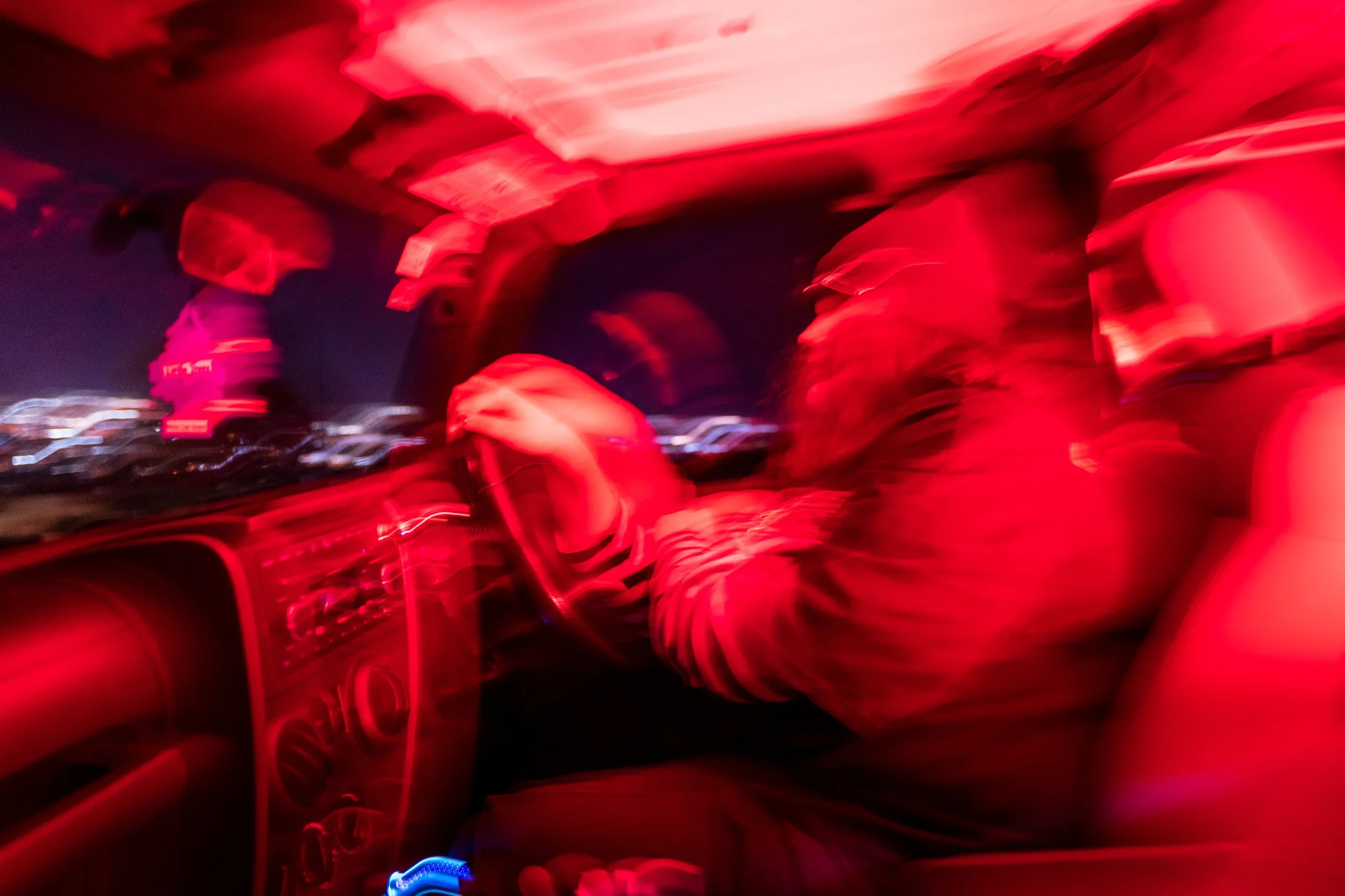 A blurry photo taken inside a car at night, showing a person wearing a helmet and dark clothing driving, with city lights visible through the window, illuminated predominantly in red.