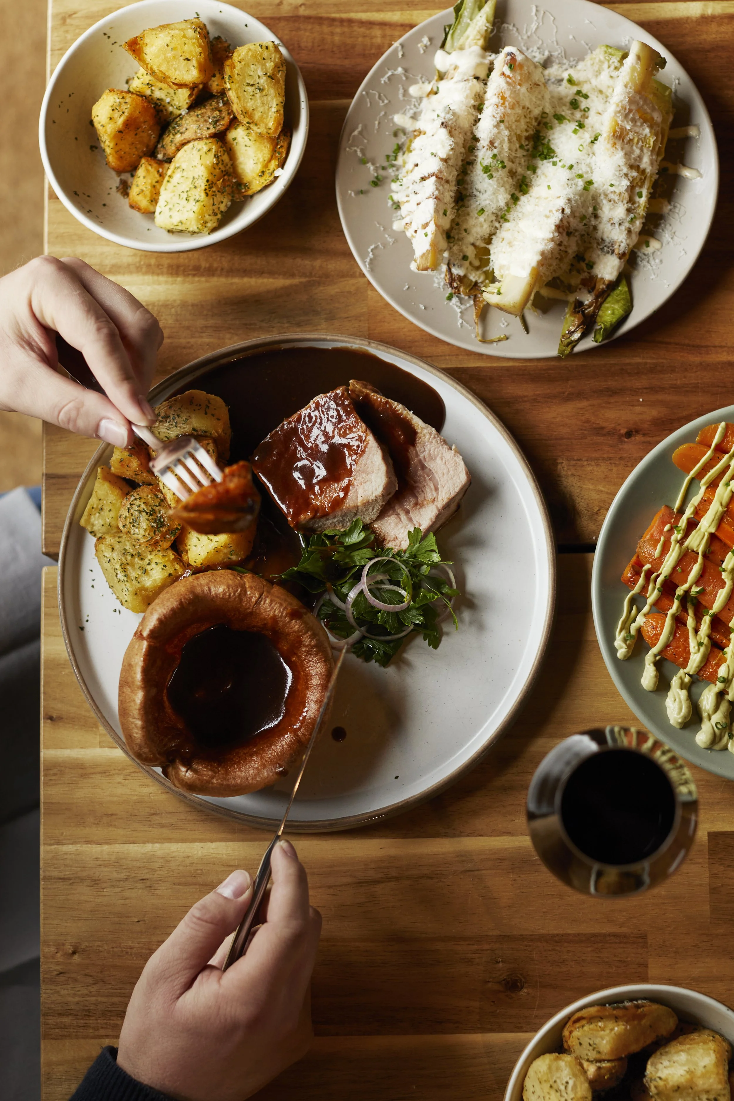 A table with American-style food including roasted potatoes, a plate of baked cauliflower with cheese, a plate of BBQ pork with potatoes, a salad with carrots and avocado, and a glass of dark beer.