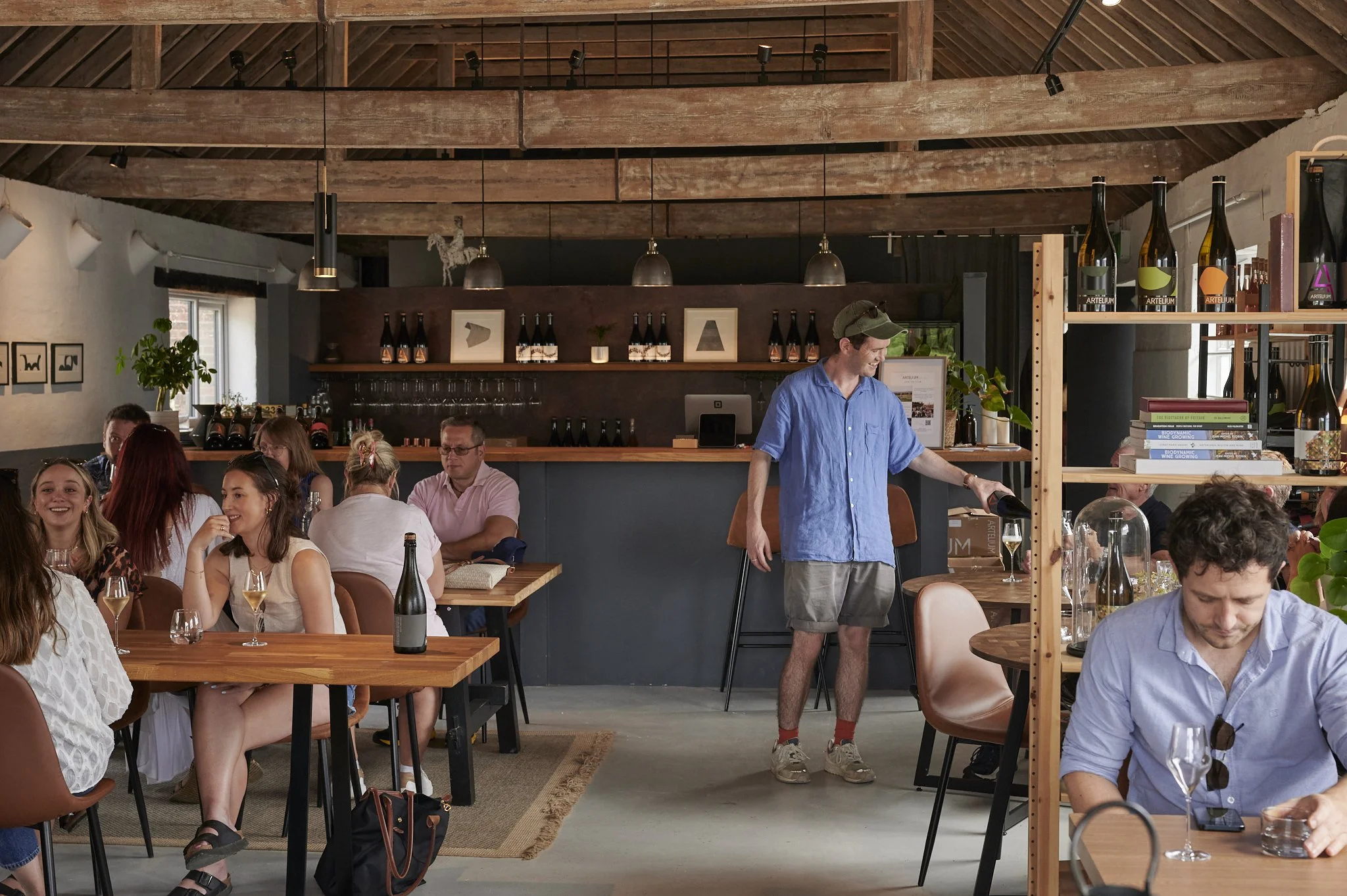 Indoor restaurant scene with several people sitting at tables enjoying drinks, a bartender pouring wine, and a warm wood interior with shelves and plants.