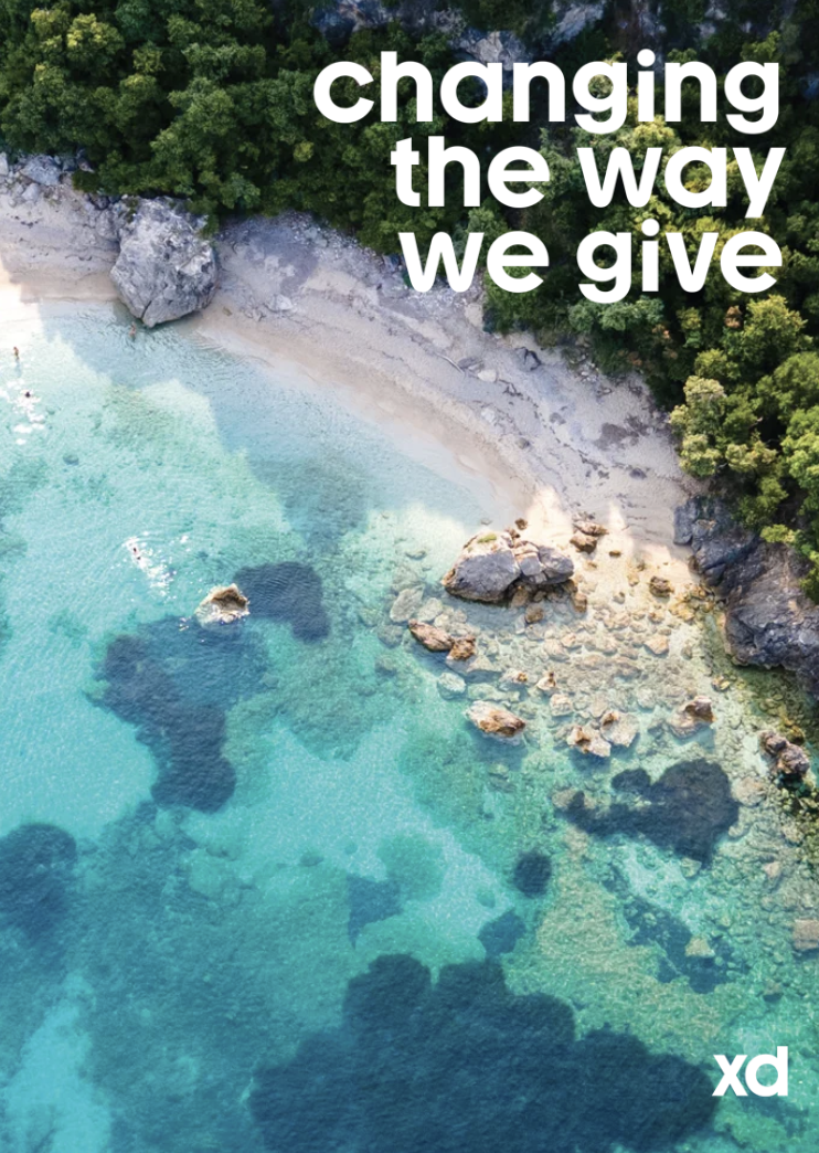 Aerial view of a rocky shoreline with clear turquoise water and lush green trees in the background, with the text 'changing the way we give' overlaid.