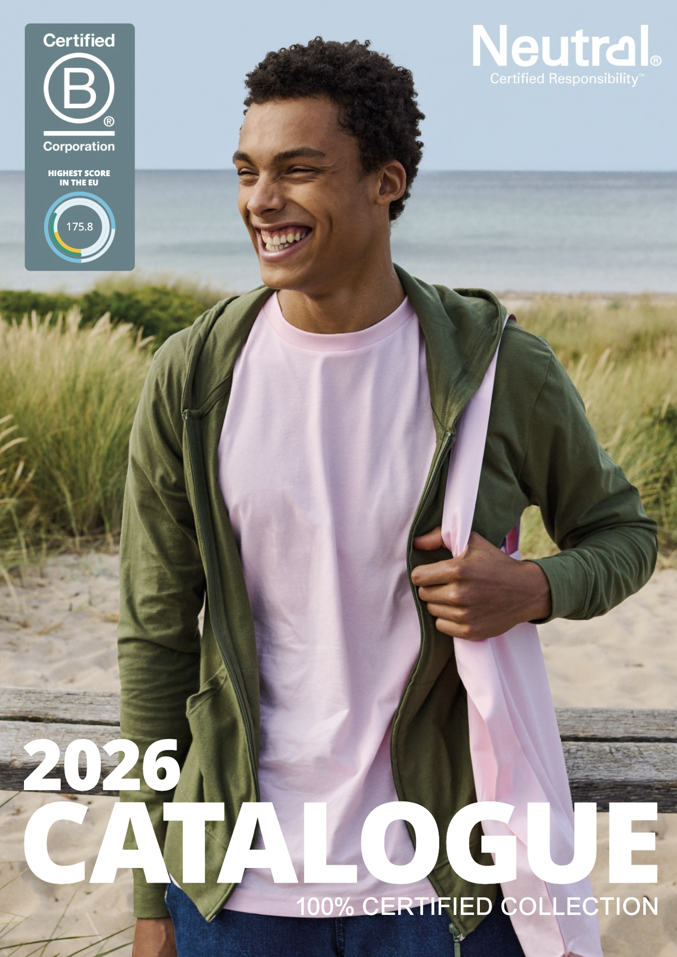 A smiling young man with curly hair wearing a pink shirt and olive green jacket, standing outdoors near a beach with tall grass and ocean in the background.