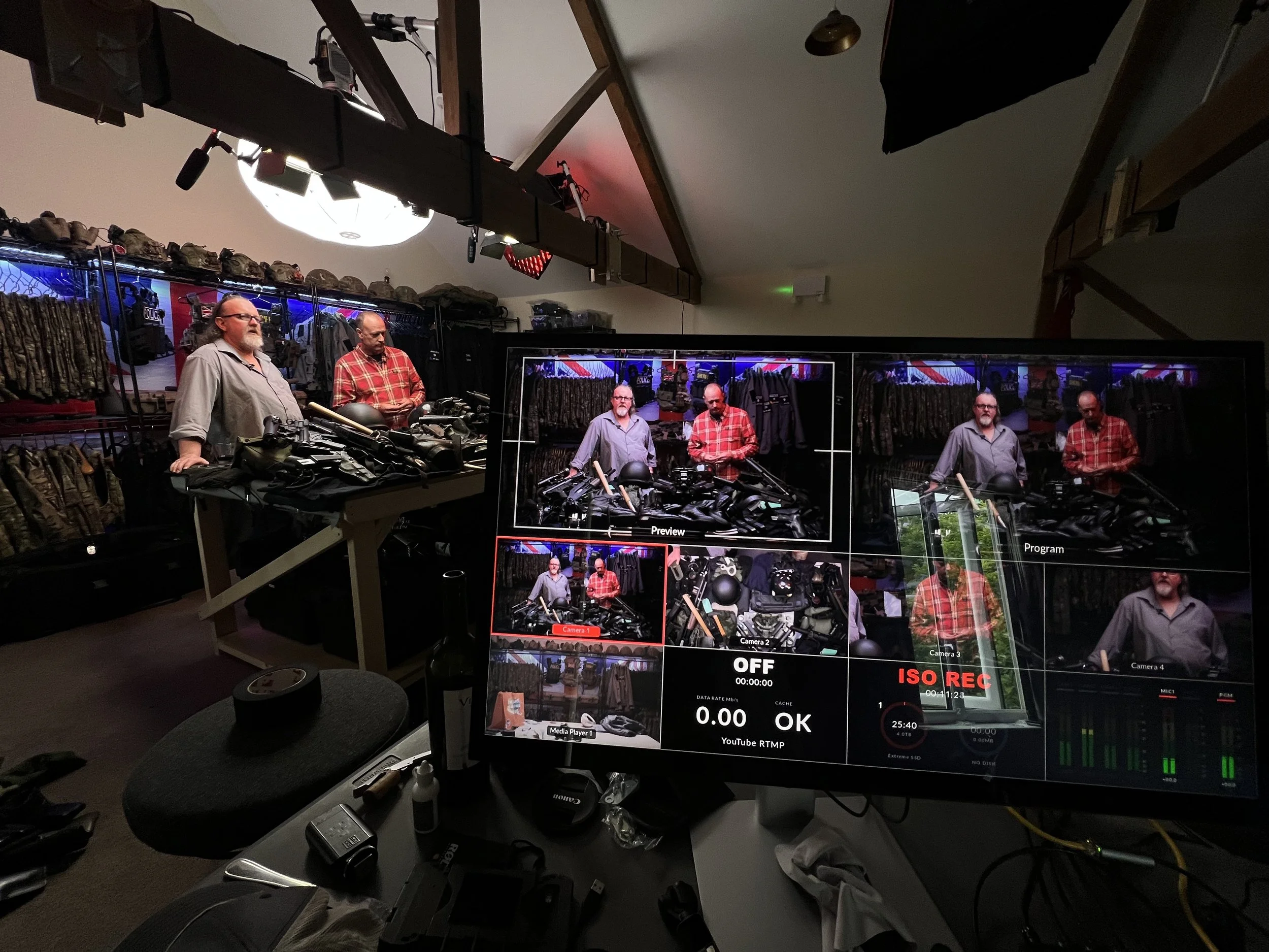 Two men standing behind a table with firearms and tactical gear, with a camera monitor in the foreground displaying a live view of the scene.