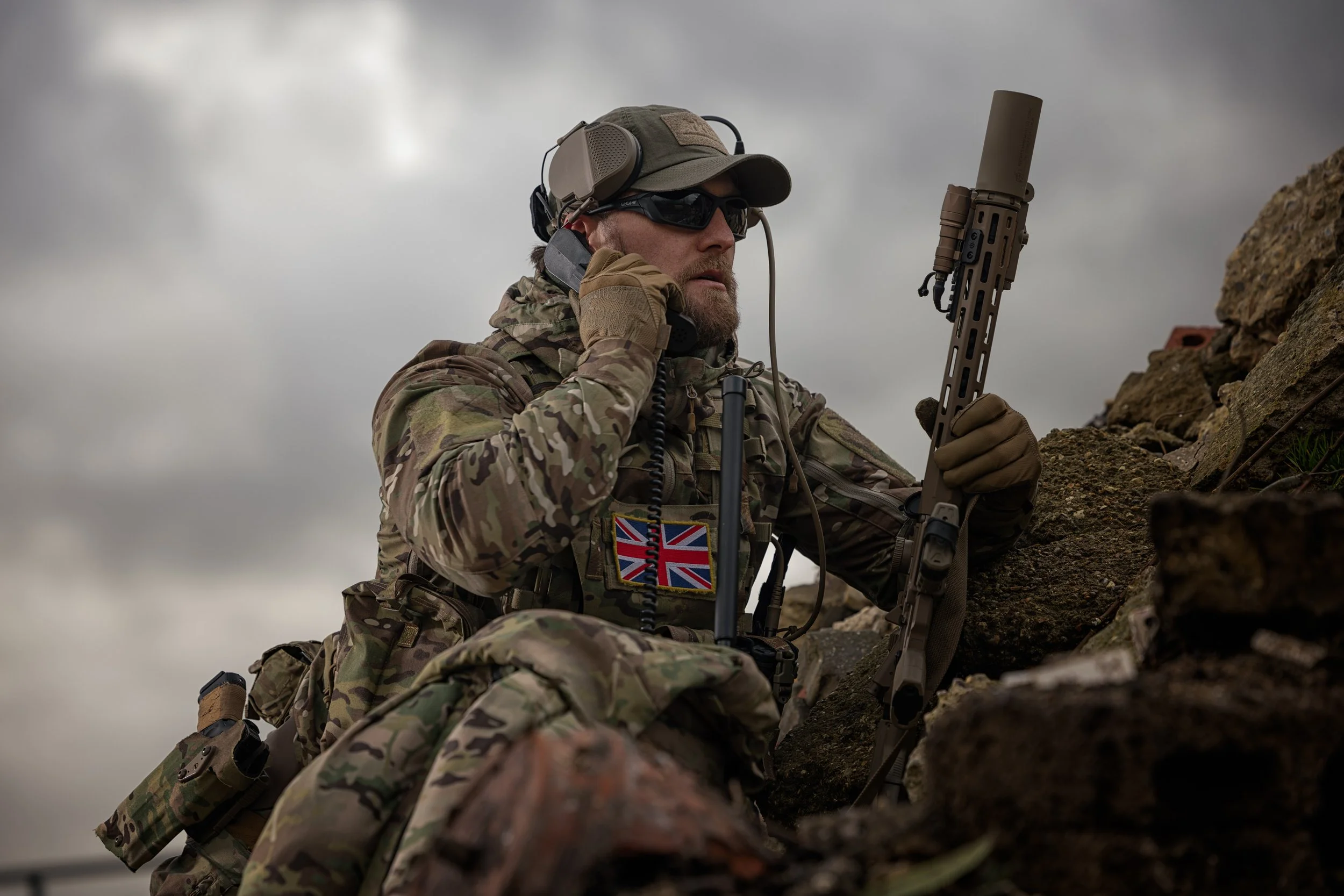 A soldier in camouflage gear climbing over rocks, talking on a radio, holding a rifle with a suppressor, and wearing sunglasses and a cap. The background shows a cloudy sky.