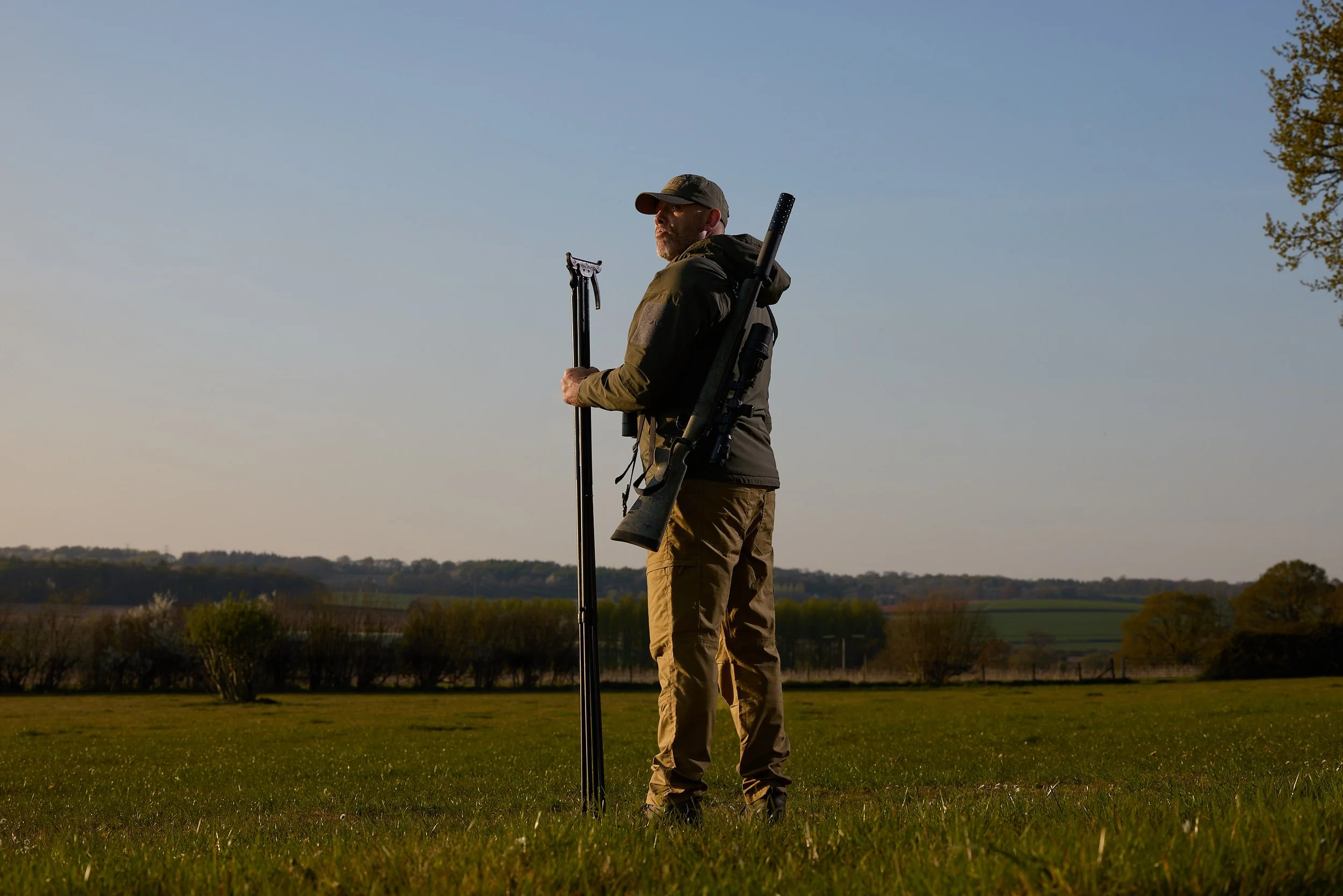 A man in outdoor gear standing in a grassy field holding a tripod, with a rifle or similar object slung over his shoulder, looking into the distance during sunset or sunrise.