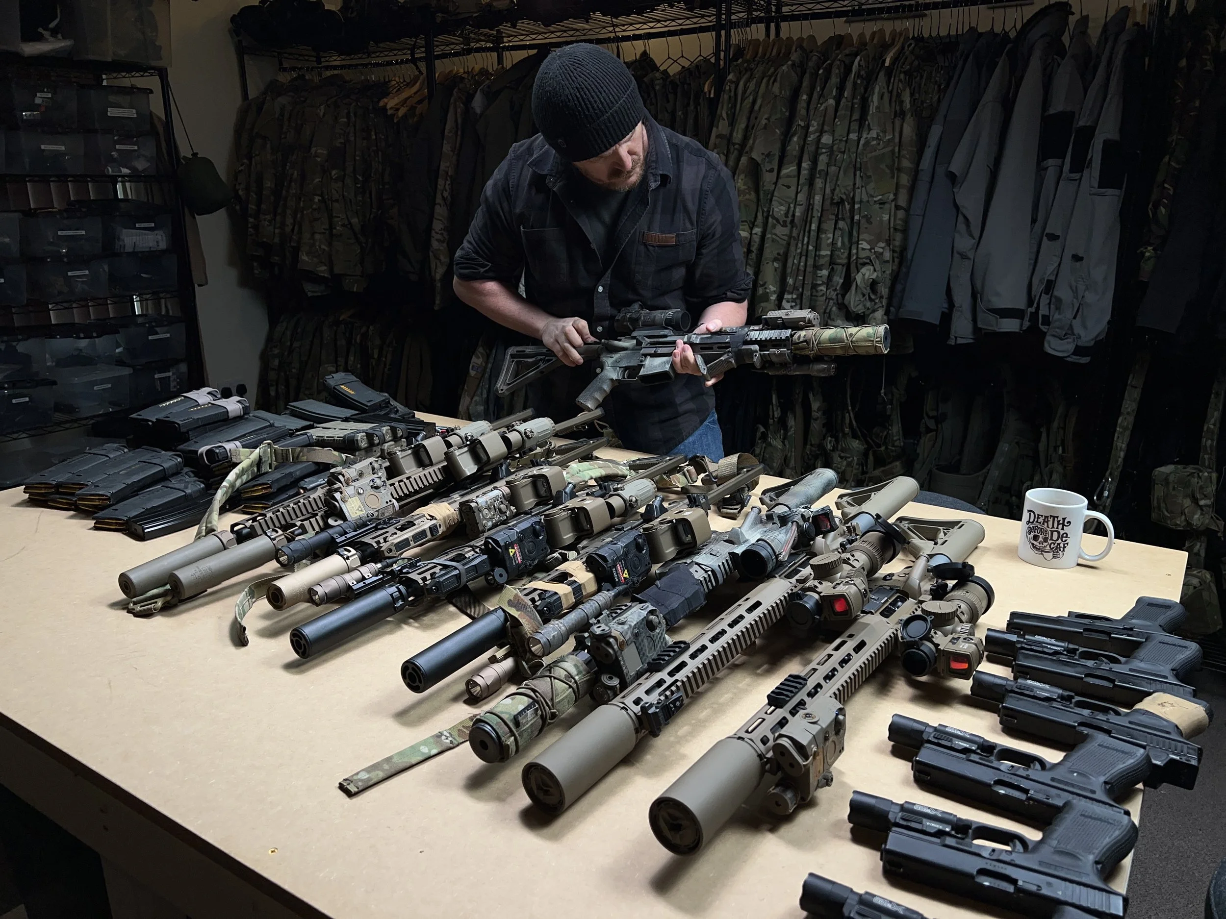 A person inspecting a rifle in a room with multiple firearms, tactical gear, and camouflage clothing on the table and background.