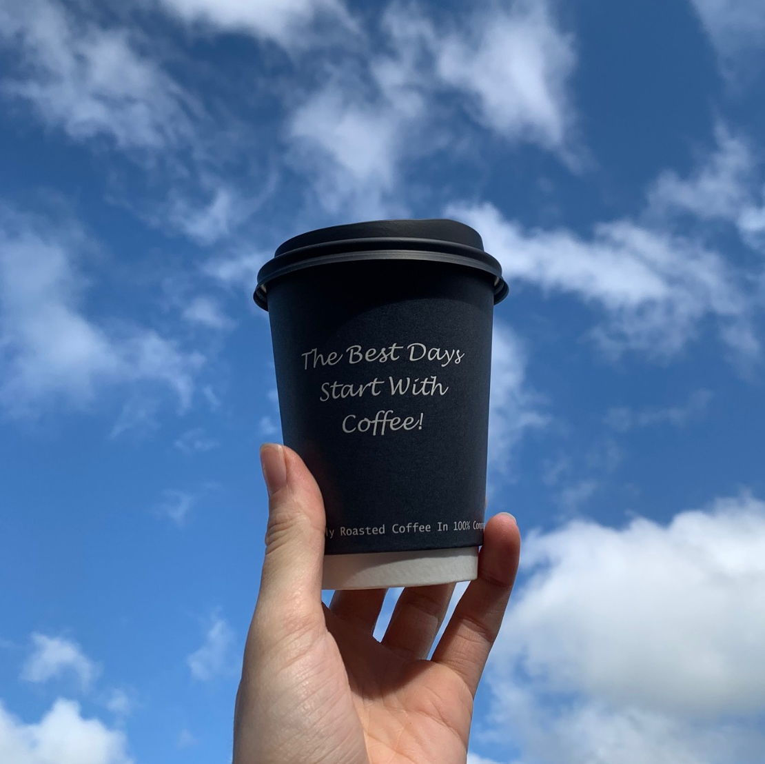 Hand holding a black coffee cup outdoors against a blue sky with scattered clouds. The cup has the words "The Best Days Start With Coffee!" printed on it.