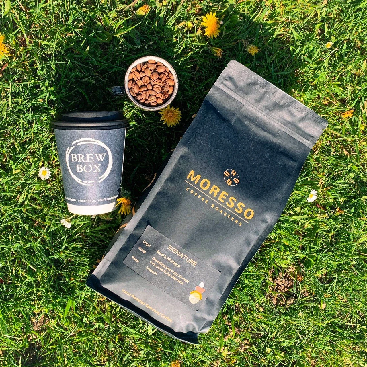Coffee beans in a small cup, a black paper coffee bag labeled Noresso Coffee Roasters, and a black coffee cup sleeve with Brew Box logo, all placed on green grass with yellow and white flowers.