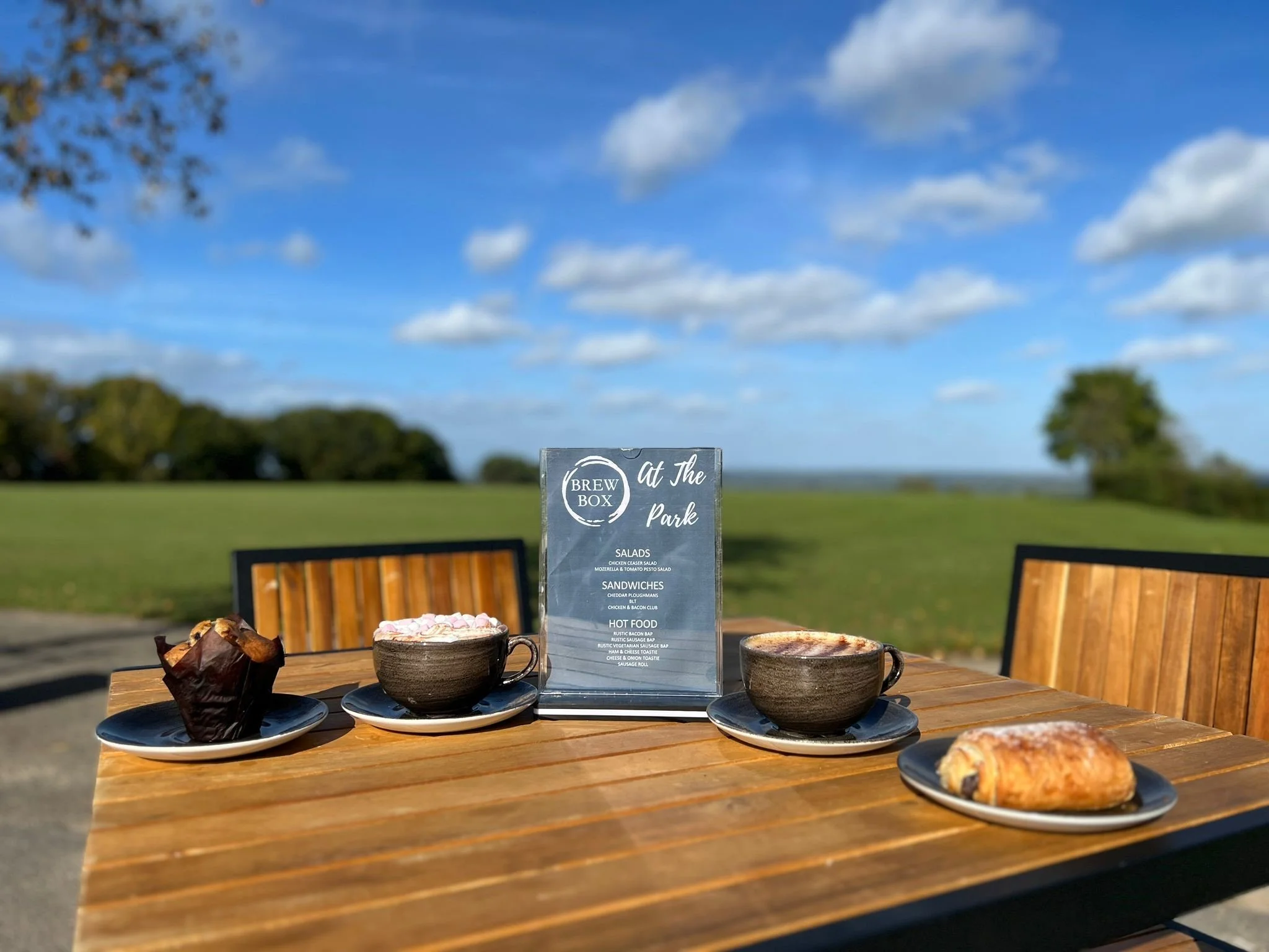 Outdoor table with coffee cups, baked goods, and a menu sign at a cafe, with trees and a blue sky with clouds in the background.