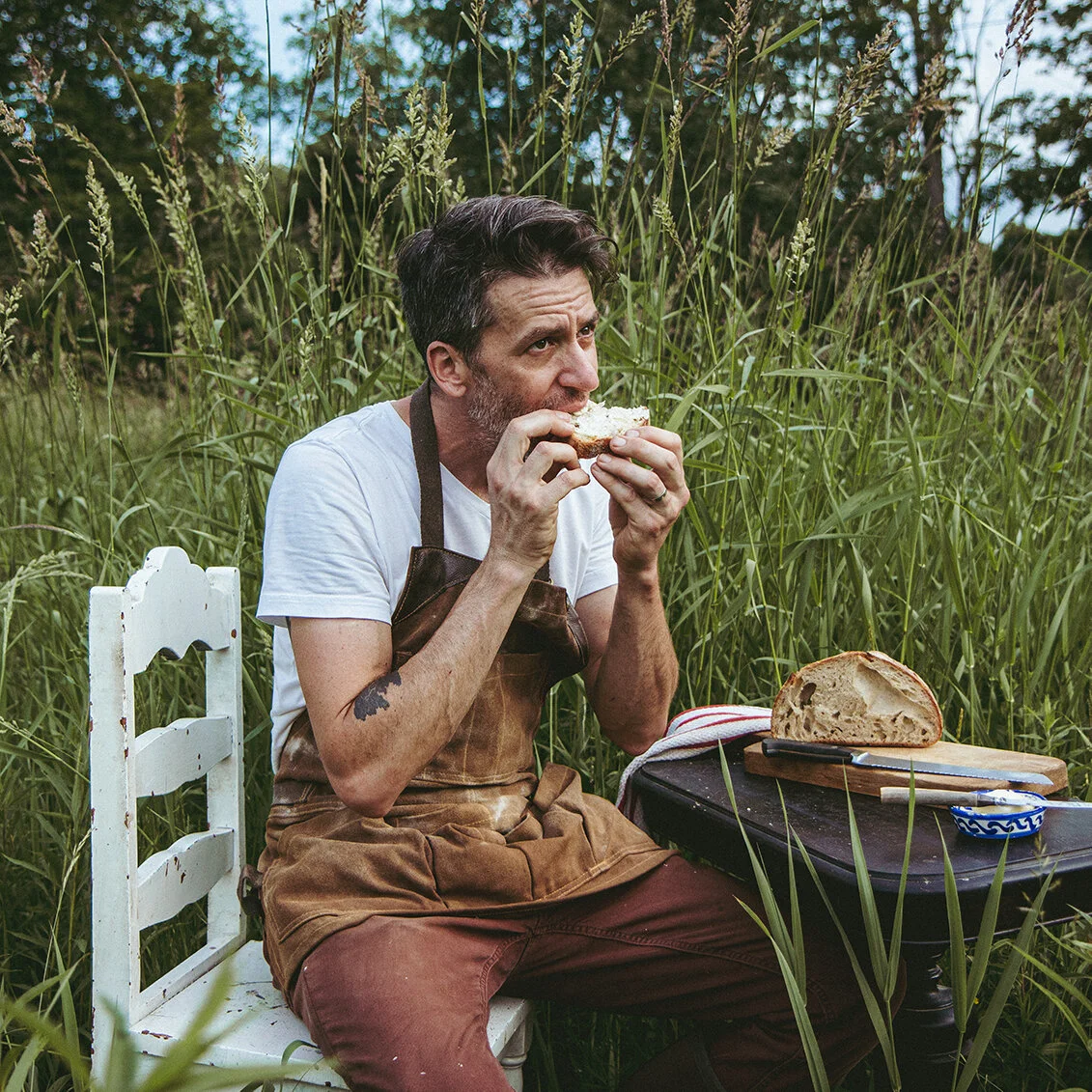 Brian is sitting on a white chair in a field with a table of bread in front of him as he's eating a piece of bread with butter