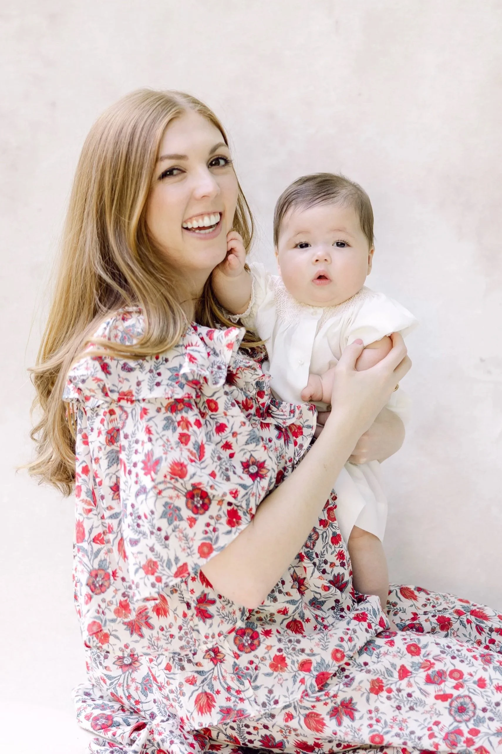 A woman with long, light brown hair, wearing a floral dress, holding a baby with dark hair and a white dress, against a plain light-colored background.