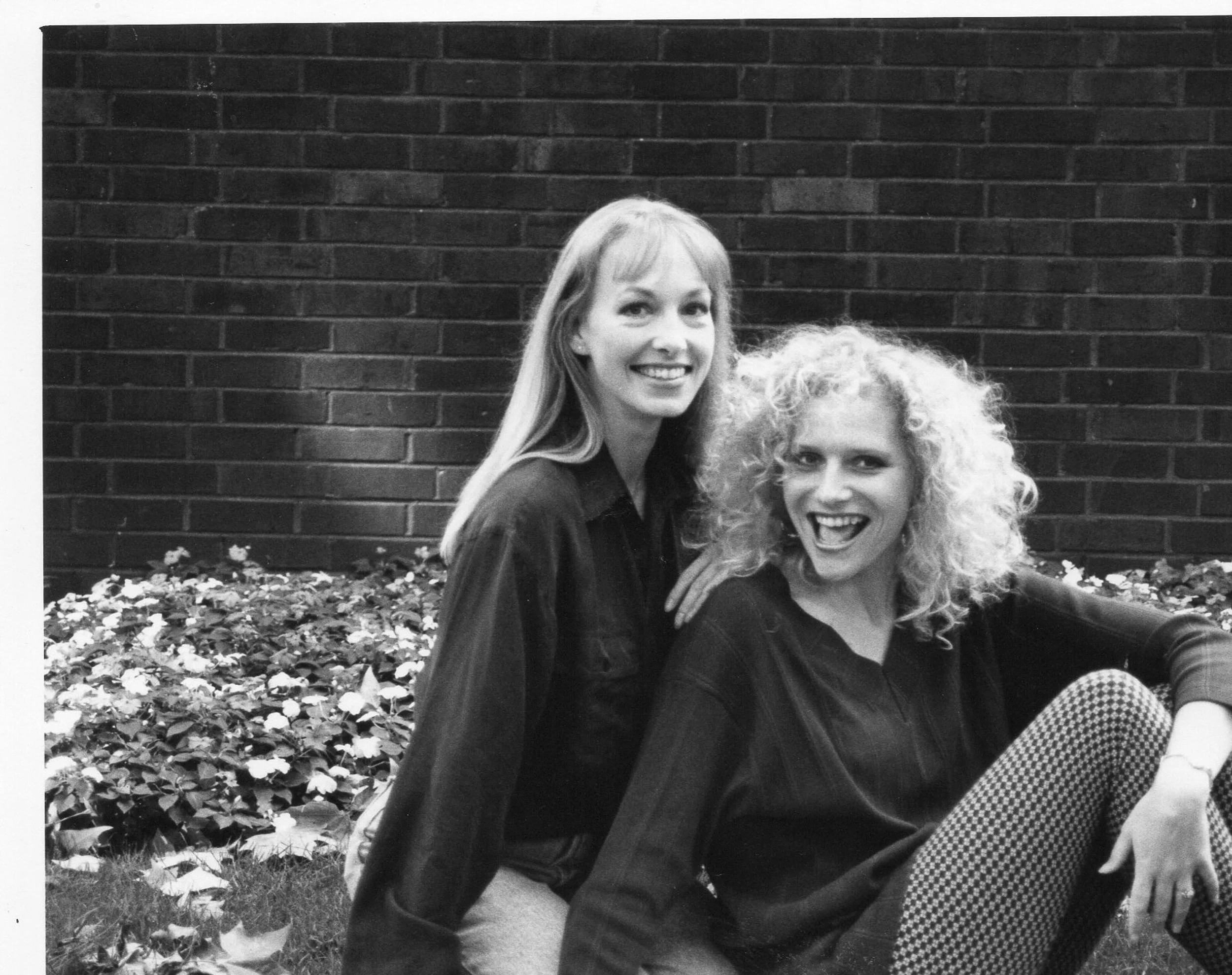Founders Caroline with long curly blond hair and Lyndsey with long straight hair smiling wide, with a brick wall behind.