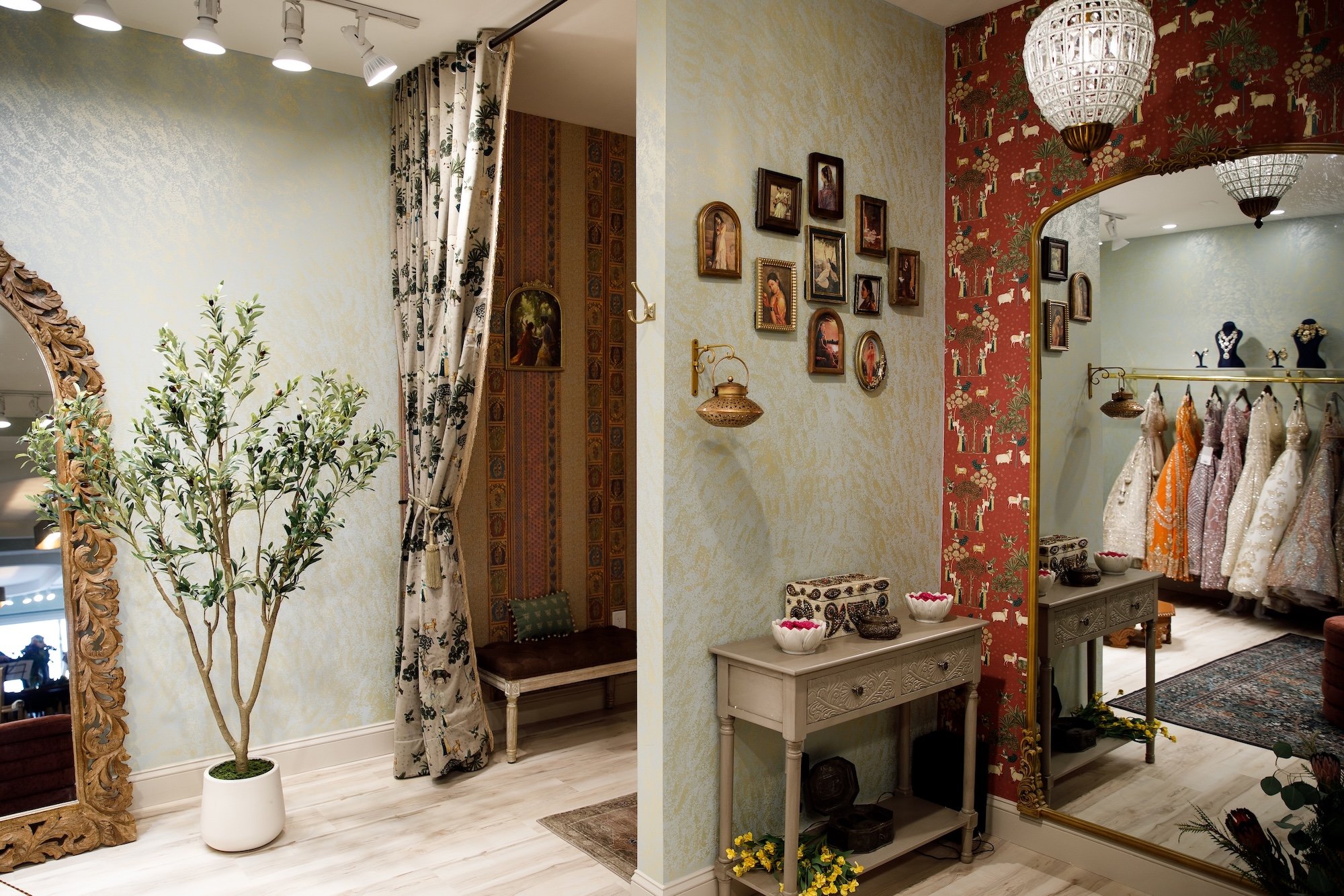 Interior view of a boutique shop with vintage decor, including a large ornate mirror, a potted plant, a wall with framed pictures, a small table with decorative bowls, a clothing rack with dresses, and hanging chandeliers.